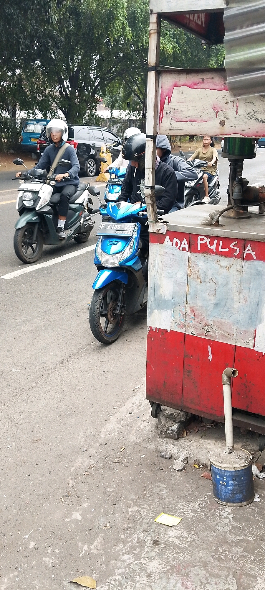 A street scene in Depok, Indonesia, captures several people on motorbikes near a roadside stall. The foreground is dominated by a red and white cart, likely a food or goods vendor, with the partial text "ADA PULSA" visible on its side, indicating a mobile credit service. A blue and white bucket, possibly containing a drain pipe, rests near the cart.
On the street, a person wearing a black hooded sweatshirt and a black helmet rides a blue motorbike. They are smoking a cigarette. To their left, another motorbike, green and white, is ridden by a person in a blue sweater and white helmet, carrying a beige strap over their shoulder. Further back, a woman on a motorbike, wearing a light brown shirt, is visible.
The background shows trees and a black SUV parked on the side of the road. The asphalt surface of the road appears worn, with some debris scattered around the foreground. The overall impression is a typical urban street environment in Indonesia, with daily life activities unfolding around roadside commerce.