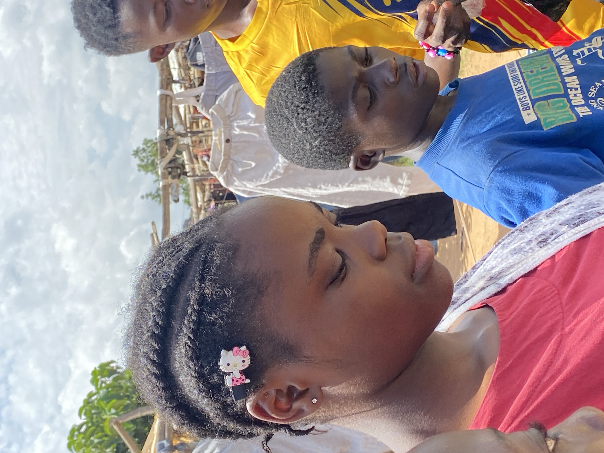 The image shows an outdoor scene with at least three individuals. The main subject is a girl in the foreground, wearing a red shirt, with braided hair adorned with a Hello Kitty hair clip. She appears to be resting or sleeping. Behind her, partially visible, is a boy in a blue t-shirt and further back is another person in a yellow shirt. The background reveals elements suggesting an outdoor market or public gathering, with indeterminate objects and structures. The location is Riyom, Nigeria.