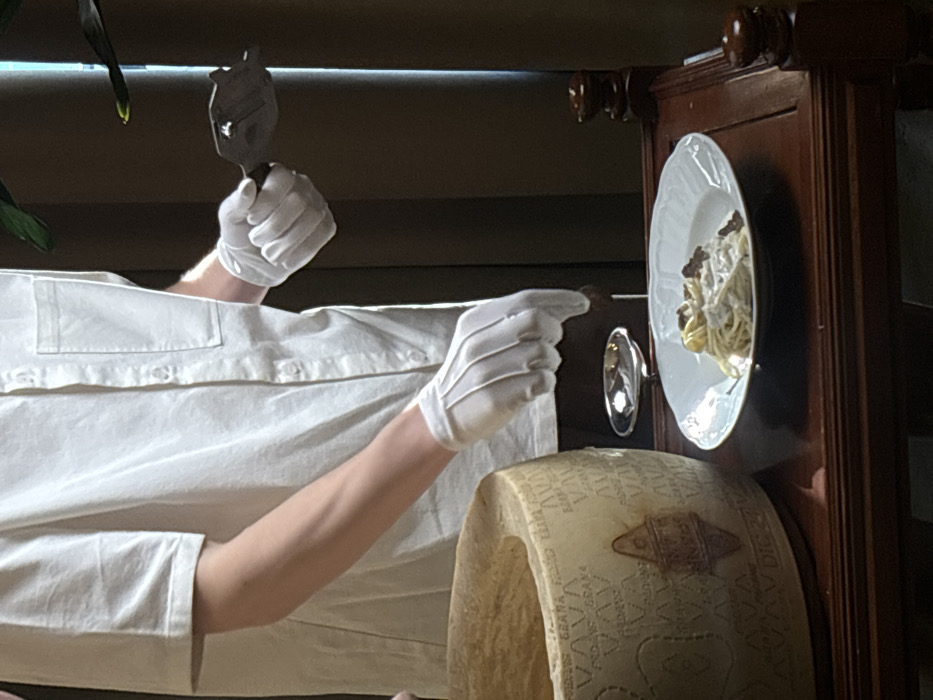 A person, identifiable by their torso and arms, is depicted wearing a white short-sleeved button-down shirt with a left chest pocket and white gloves on both hands. The individual's right hand holds a dark metal truffle shaver, from which a dark, small object (presumably a truffle) is being dispensed onto a white ceramic plate. This plate contains spaghetti pasta coated in a creamy white sauce, with dark fragments already present. The left hand is positioned over a small, shiny silver bowl located on the table to the right of the plate. To the left, a large, partially hollowed wheel of Grana Padano cheese is visible, bearing its characteristic rind markings. All objects are situated on a dark brown wooden table featuring ornate carvings. The background is dark and out of focus, with a partial view of a green plant leaf in the upper right. The scene depicts a tableside food service within a dining establishment in Saint Petersburg, Russia.