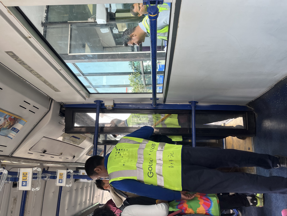 The image displays the interior of a public bus during daytime, likely in Guwahati, India. The primary subject is a man standing in the aisle on the right side of the frame, seen from behind and slightly to his left. He wears a long-sleeved blue shirt under a bright yellow high-visibility vest. The vest has text printed on the back, which appears to read "Gound force" (possibly "Groundforce"). Reflections in the glass partition behind him show another man, likely a colleague, also in a yellow vest, and a blurred reflection of another person's face in the upper left section of the glass. Part of a person or child wearing colorful clothing is partially visible below the main subject.
The bus interior features a blue floor, light grey walls and ceiling panels, and blue support poles. Transparent plastic handles with "LIC" (Life Insurance Corporation) logos are attached to a rail on the left. On an overhead ceiling panel, the text "ASHOK LEYLAND" is prominently visible, indicating the bus manufacturer. Vents are also present on the ceiling.
Through the windows and reflections, green foliage from trees or bushes, parts of urban buildings, and a bright, clear sky are visible. The scene captures a typical public transport environment, with the man in the vest likely serving as a conductor or staff member.
