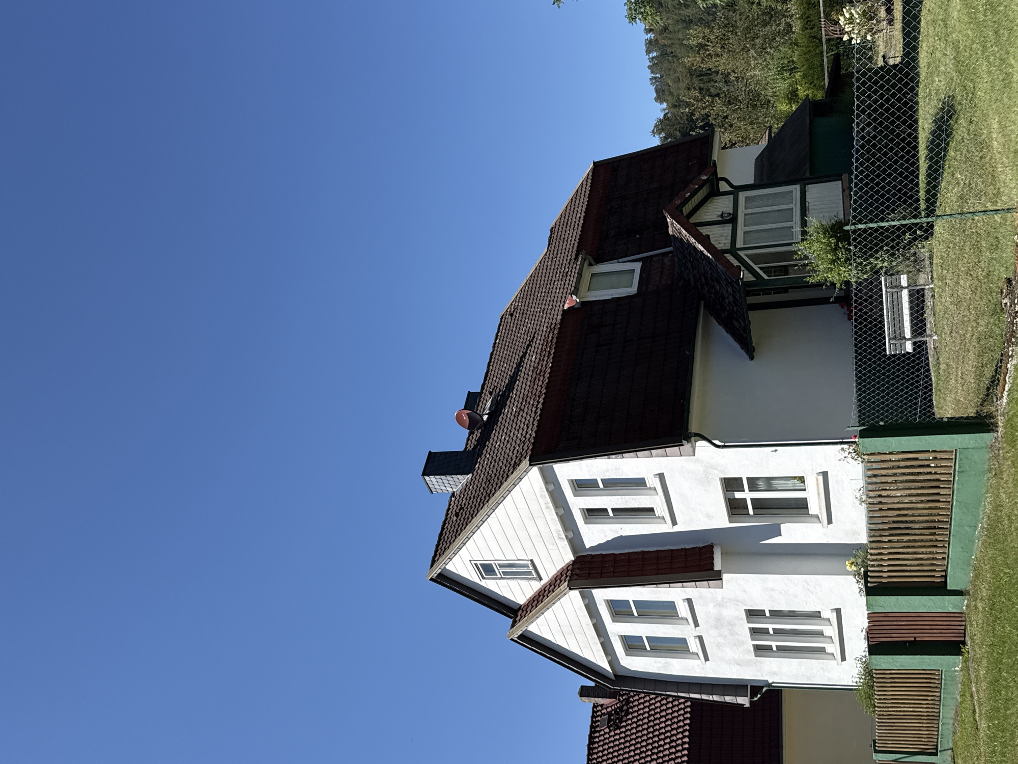 The image depicts a two-story white house with a dark red tiled roof, situated in the city of Bad Lauterberg im Harz, Germany. The house has multiple windows with white frames. A portion of an adjacent house, also with a similar tiled roof, is visible to the left. In front of the house, there is a wooden fence with green painted posts and a small wooden gate. Beyond the fence, a chain-link fence encloses a grassy area with a white bench. The background features trees and a clear blue sky. The scene appears to be a residential area on a sunny day.