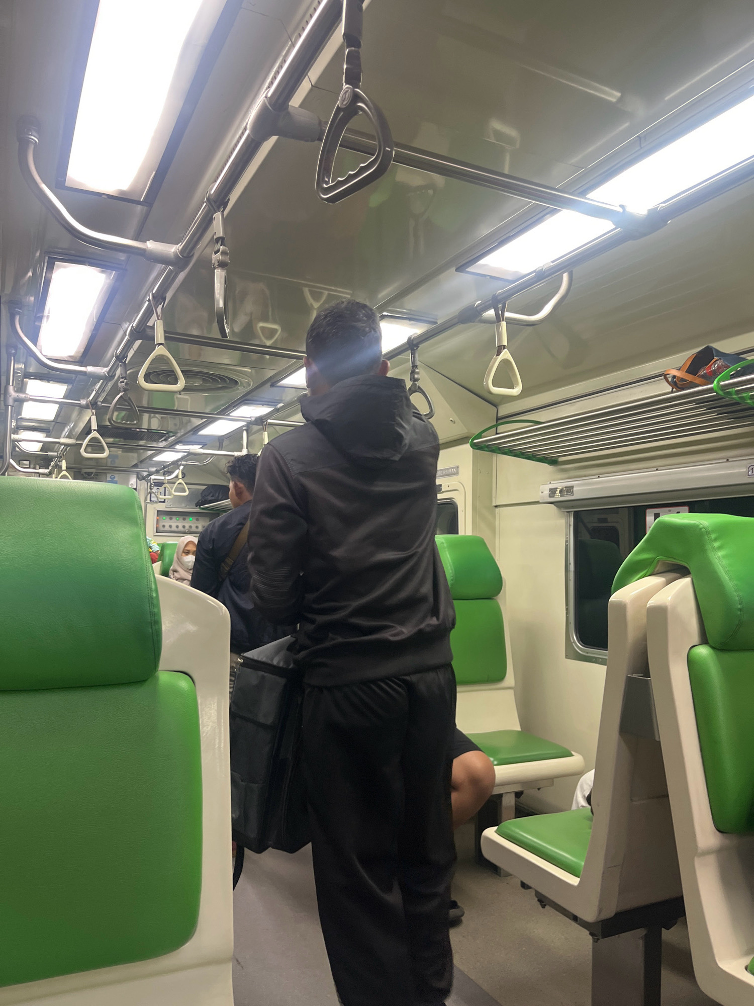 The image depicts the interior of a train carriage, likely in Babat, Indonesia, given the context. The perspective is from behind a person wearing a black hoodie and black pants, who is facing forward into the carriage. The carriage features rows of bright green seats with white accents. Overhead, there are handrails and luggage racks. The lighting appears to be artificial, coming from long fluorescent fixtures on the ceiling. Another person, also wearing dark clothing, is visible in the mid-ground, and a person wearing a headscarf is seated further down the aisle. The overall impression is one of a typical public transportation environment.