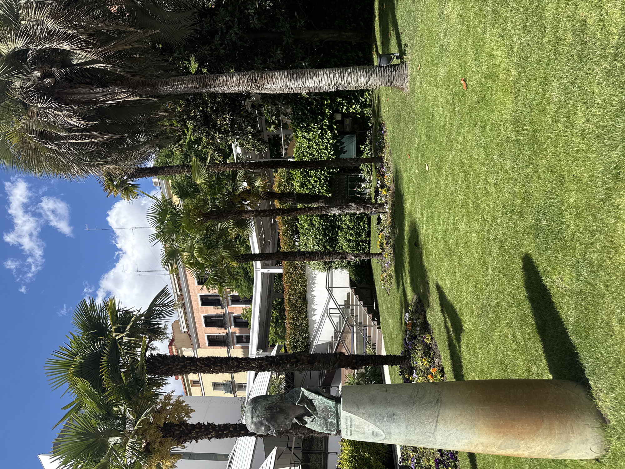 This is an outdoor scene in Madrid, Spain, featuring a park or garden setting. The main subjects include several palm trees, a grassy lawn, and a bronze bust statue on a cylindrical pedestal. The sky is visible with a mix of blue and white clouds. Buildings are partially visible in the background. The statue is positioned near the front of the frame, surrounded by flowers. The grass is neatly manicured, and there's some foliage and greenery along the edges. The presence of palm trees suggests a warm, sunny climate. The lighting is bright, casting shadows from the trees and statue onto the lawn. No people are visible in the shot.