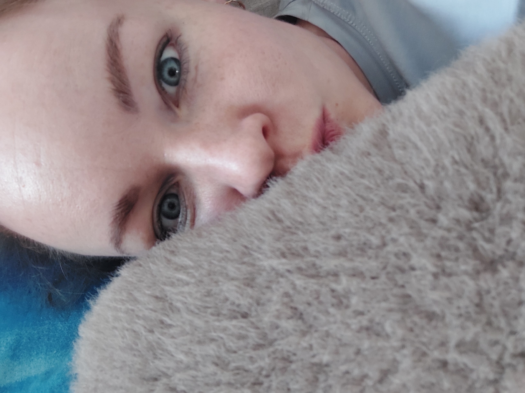A close-up, low-angle shot depicts a fair-skinned person with light eyebrows and blue eyes, lying down. Part of their face is obscured by a beige, textured, possibly fuzzy material, that may be a sweater or blanket. Hints of blue, possibly fabric, appear on the left. A gray shirt is seen in the upper right. The location is unidentified as the city of Chaikovsky, Russia.