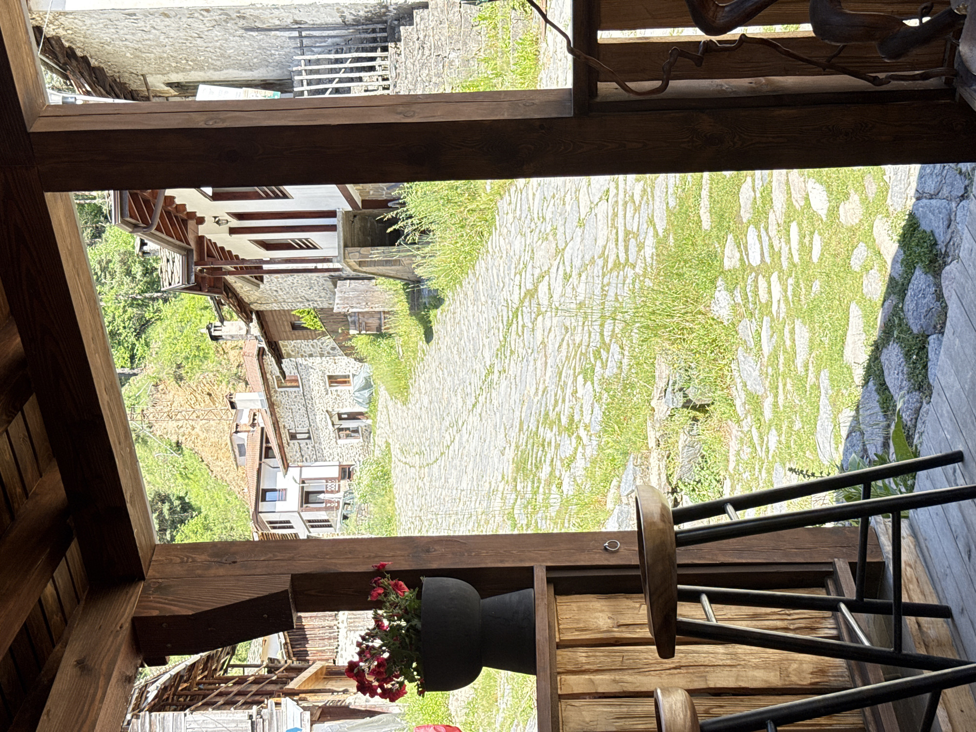 The image shows a view from a wooden veranda in the village of Shiroka Laka, Bulgaria. In the foreground, two wooden bar stools are visible on a wooden deck. To the left, a dark planter filled with red flowers sits on a wooden shelf. Beyond the veranda, a cobblestone street winds uphill, lined with traditional stone houses. Green vegetation, including grass and trees, grows alongside the street and on the surrounding hillsides. The architecture suggests a historic Balkan village setting.