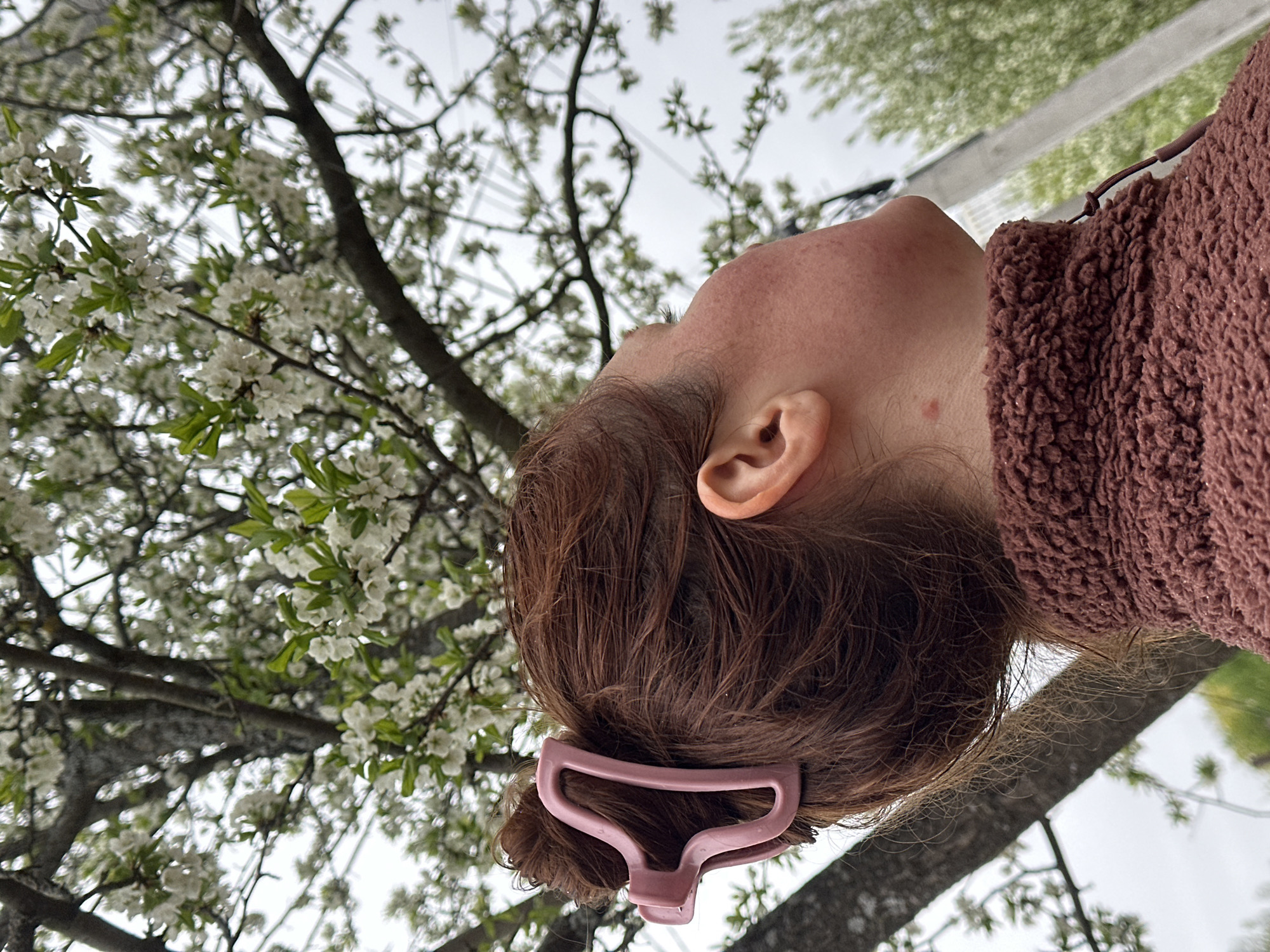 The image shows a person looking up at a blooming tree. The person has reddish-brown hair pulled back with a pink hair clip. The tree is filled with white blossoms. The scene appears to be outdoors. It is not possible to determine the location in Nizhny Novgorod, Russia from the image.