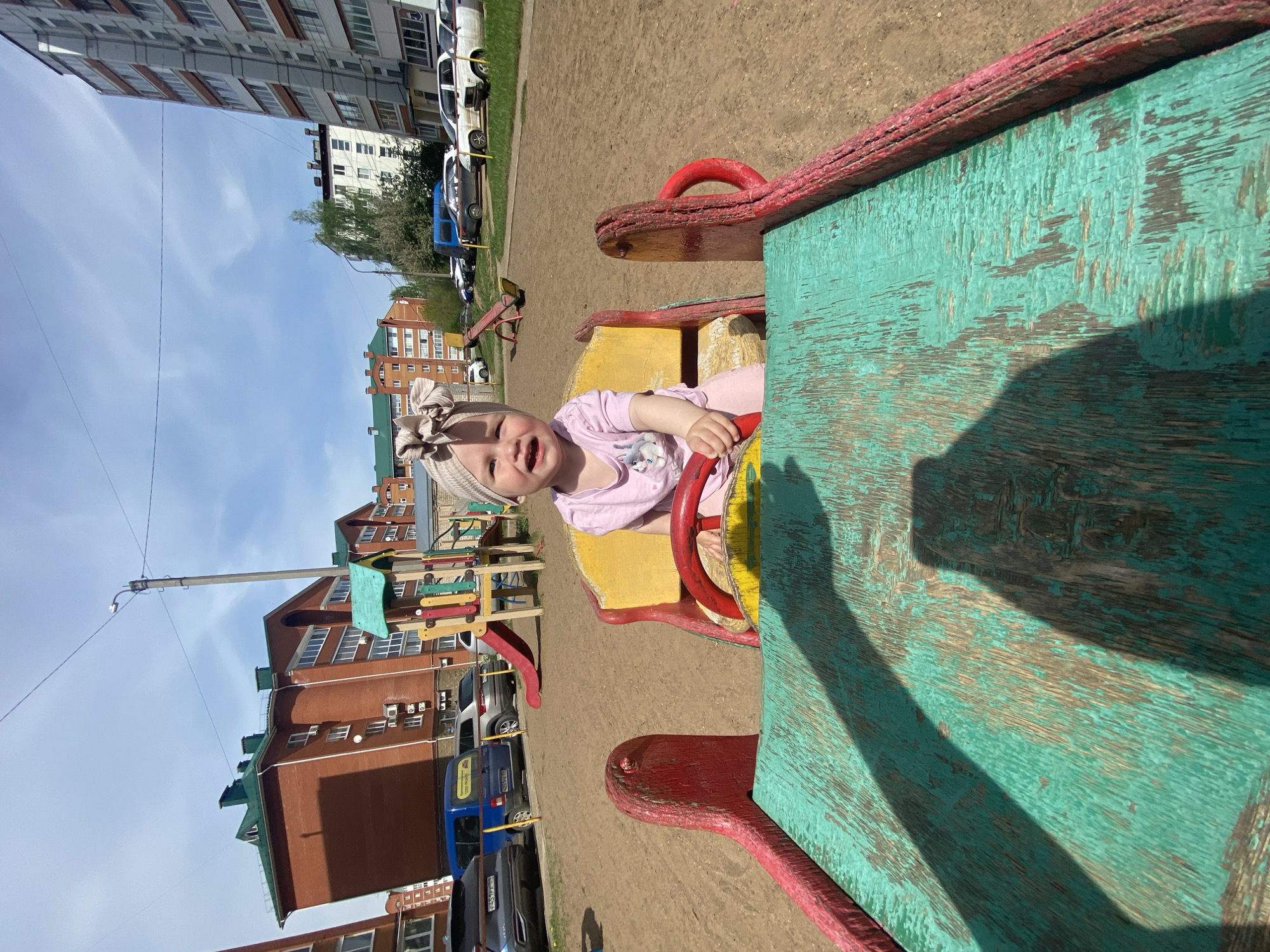 A young child, wearing a light pink shirt and a beige headband with a bow, is sitting on a yellow and red playground ride with a weathered turquoise top at what appears to be a playground in Chaikovsky, Russia. The child is looking at the camera. Behind the child are several buildings of various colors including brick and cream. Parked cars are visible on the street adjacent to the playground. The sky is pale blue with some white clouds. A streetlight can be seen in the background.