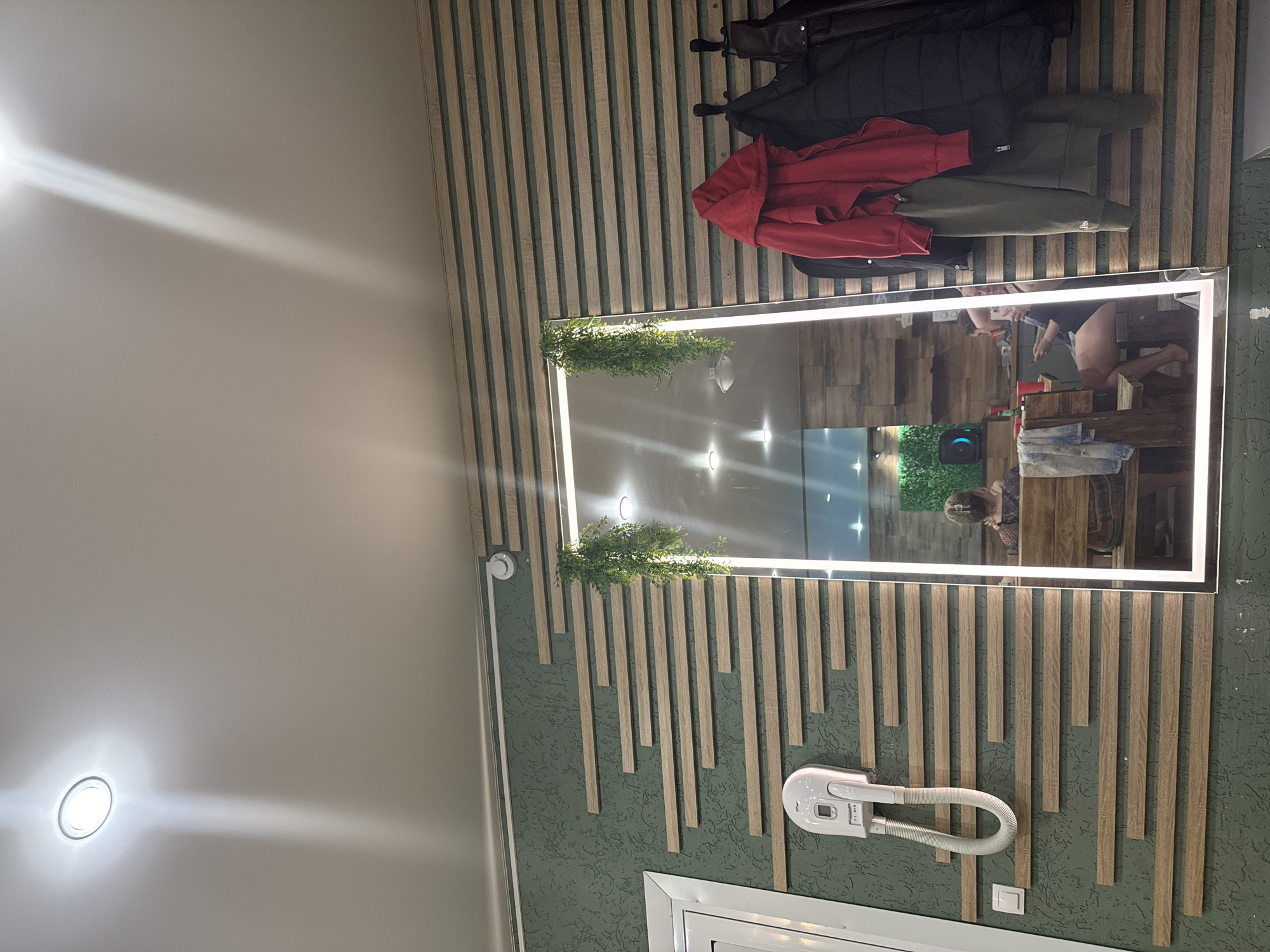 This image captures an indoor scene, likely a dressing room, salon, or entryway in Chaikovsky, Russia. The primary subject is a large, rectangular mirror with integrated LED backlighting, mounted on a wall adorned with green textured wallpaper and vertical wooden slats.
Reflected in the mirror is a partial view of a person, appearing to be a female, wearing dark shorts and a light-colored top. She seems to be looking into the mirror, possibly taking a photograph or adjusting her appearance. The reflection also shows glimpses of an interior space behind the viewer, including what appears to be a wooden surface or counter with some items. Artificial green foliage is decoratively placed on top of the mirror's frame at both ends.
Above the mirror, three coats hang on wall hooks: a black puffy jacket, a red hooded jacket, and an olive green jacket. Below the mirror, a white wall-mounted hair dryer with a coiled hose is installed. A white electrical switch or outlet is visible beneath the dryer, and a white smoke detector or sensor is mounted on the wall above the dryer, to the left of the mirror.
The ceiling to the left is plain and features a bright, recessed circular light, casting a distinct beam. The overall lighting is artificial and bright. Part of a white frame, possibly a door or window, is visible at the bottom right of the image. No distinct text is clearly legible on any objects within the frame.