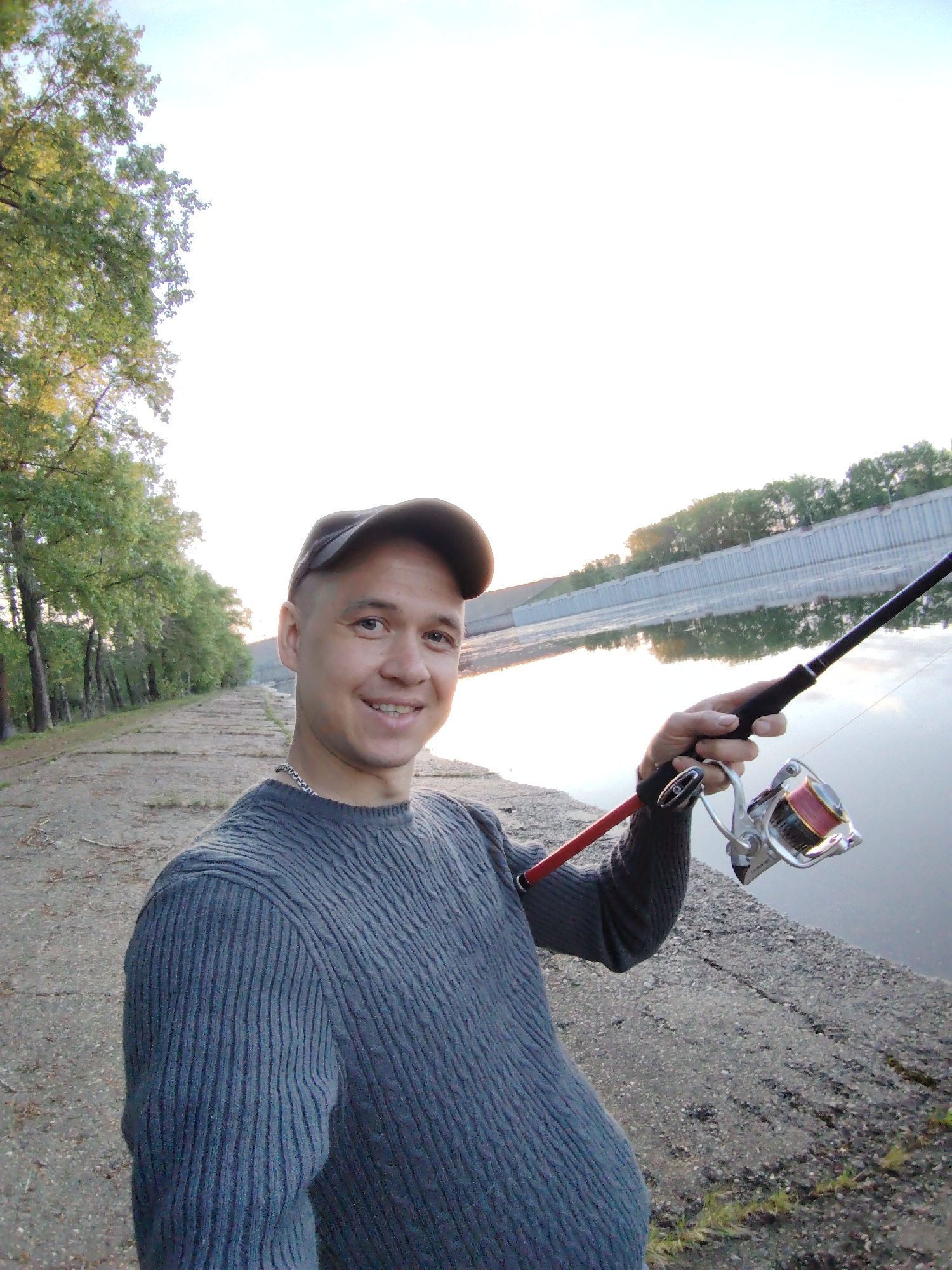 A man stands next to a body of water in Chaikovsky, Russia, holding a fishing rod. He wears a grey sweater and a baseball cap and smiles at the camera. A paved path lines the water's edge, with trees visible in the background. The water reflects the sky and nearby structures. The fishing rod extends towards the water.