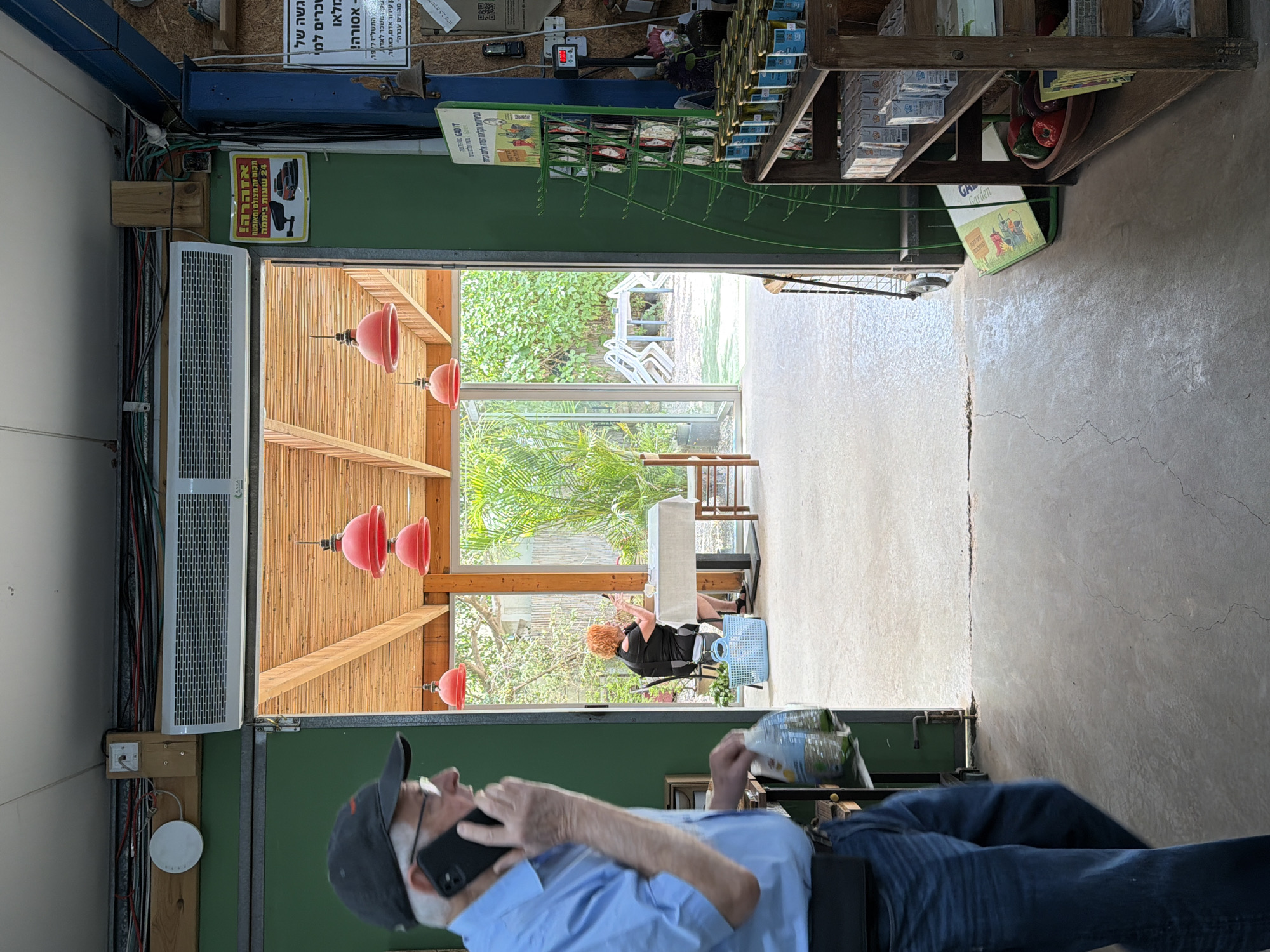 A man wearing a blue shirt and a baseball cap, holding a book and talking on a cell phone, lies on his back on what appears to be a wheeled cart in an open doorway. Through the doorway, a red-haired woman sits at a table on a porch or patio, visible through a room with a light-colored floor. The setting seems to be an interior transitioning to an exterior, possibly in Kefar HaNagid, Israel, judging by the presence of Hebrew writing on several signs and tags attached to the walls and shelving around the doorway. The room is cluttered, with shelves on the right side holding various items. The overall scene presents an indoor/outdoor transition, potentially within a retail or workshop environment.
