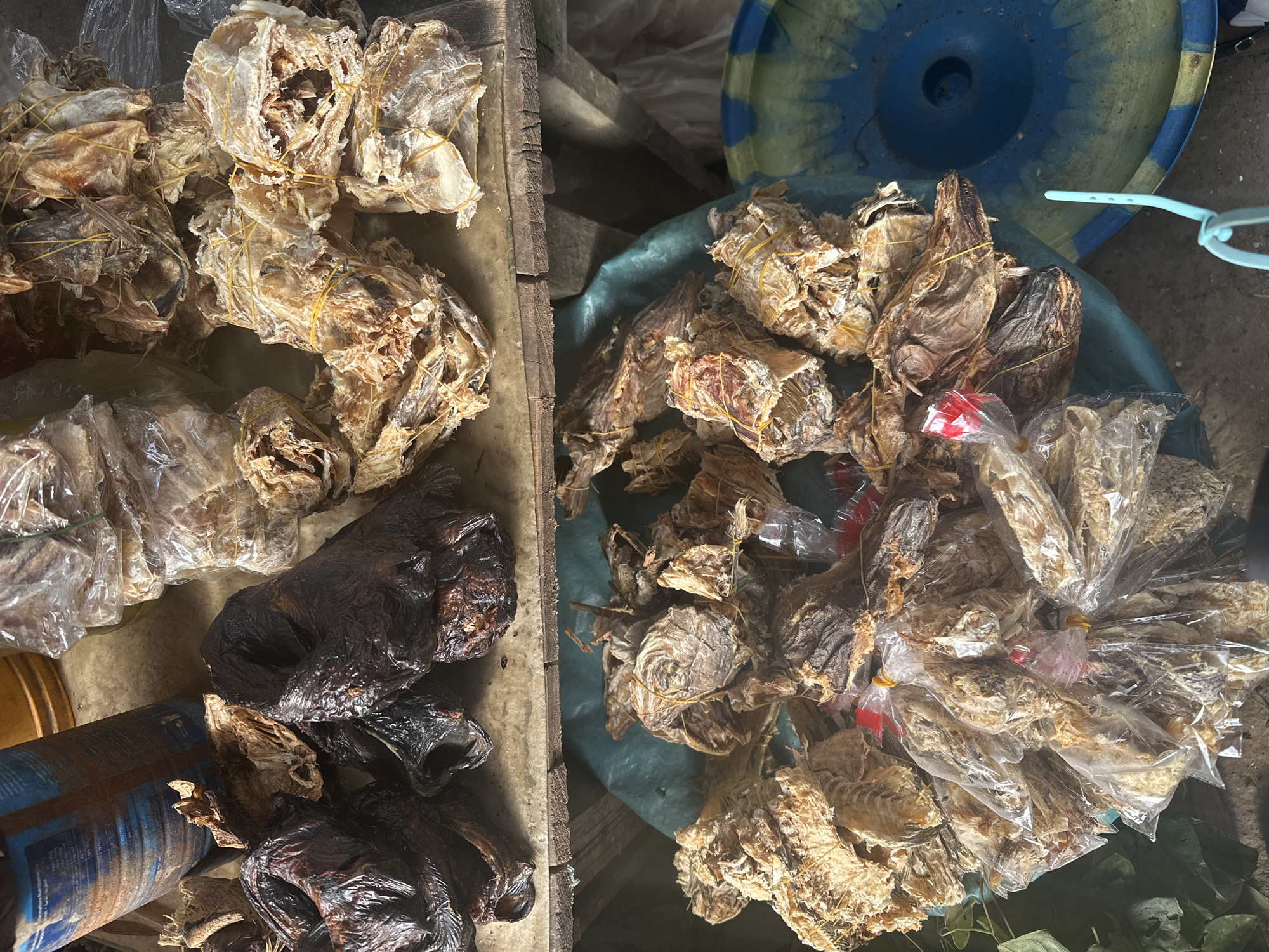 The image depicts a market stall in Lagos, Nigeria, featuring a variety of dried fish. In the foreground, a large, teal-colored plastic basin overflows with bundles of dried fish, many of which are wrapped in clear plastic bags and secured with yellow rubber bands. Some individual fish heads are visible among the bundles. To the left of the basin, a collection of darker, larger, and more irregularly shaped dried fish are displayed on a wooden surface, some appearing almost black and shiny. Behind these, on the same wooden surface, are piles of lighter-colored dried fish, also bundled and secured with yellow bands, some appearing more fragmented and flaky. In the upper left of the image, a large, dark blue and brown cylindrical object, possibly a container or barrel, is partially visible, with some text in an Arabic script on its surface. The overall scene suggests a bustling marketplace where dried fish is a prominent commodity.
