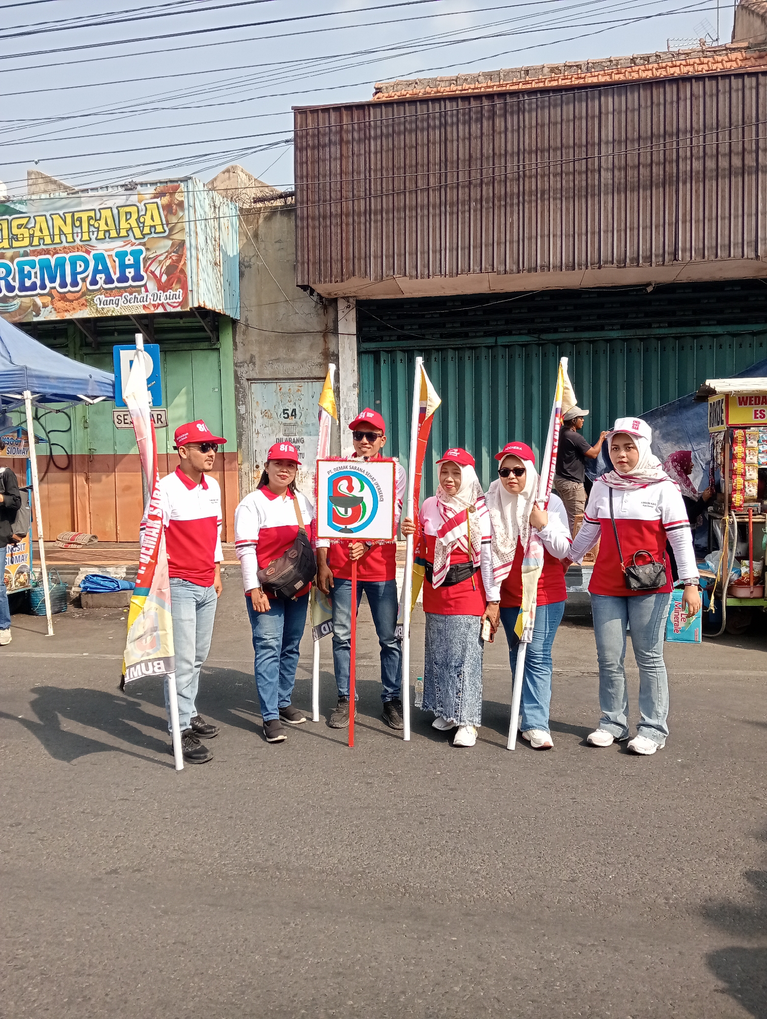 Six individuals, four women and two men, are standing in a street in Bintoro, Indonesia, posing for a photo. They are all wearing red and white shirts with the logo "81" on the chest and red caps. The men are wearing sunglasses, while the women are wearing headscarves. The group is holding flags and a sign with the same "81" logo. In the background, there are buildings with signs, one of which reads "NUSANTARA REMPAH" and "Yang Sehat Disini." There are also tents and a food cart. The ground is paved, and there are power lines overhead. The sky is clear and blue.