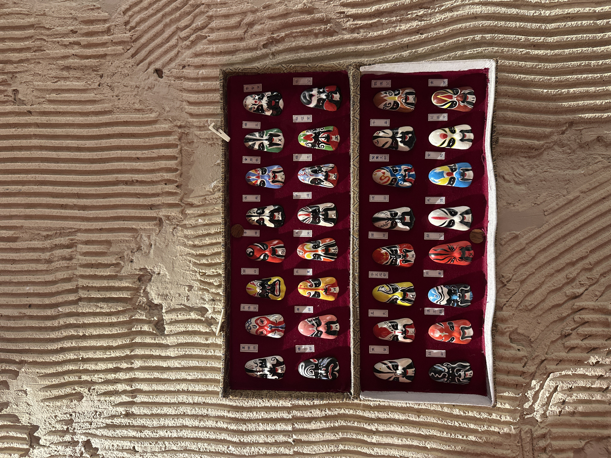 A collection of miniature Chinese opera masks is displayed in two open, velvet-lined cases mounted on a textured, beige-colored wall. Each mask is unique in color and design, with small labels in Chinese characters beside each one. The masks feature diverse facial expressions and makeup styles. The wall's texture is characterized by horizontal grooves.