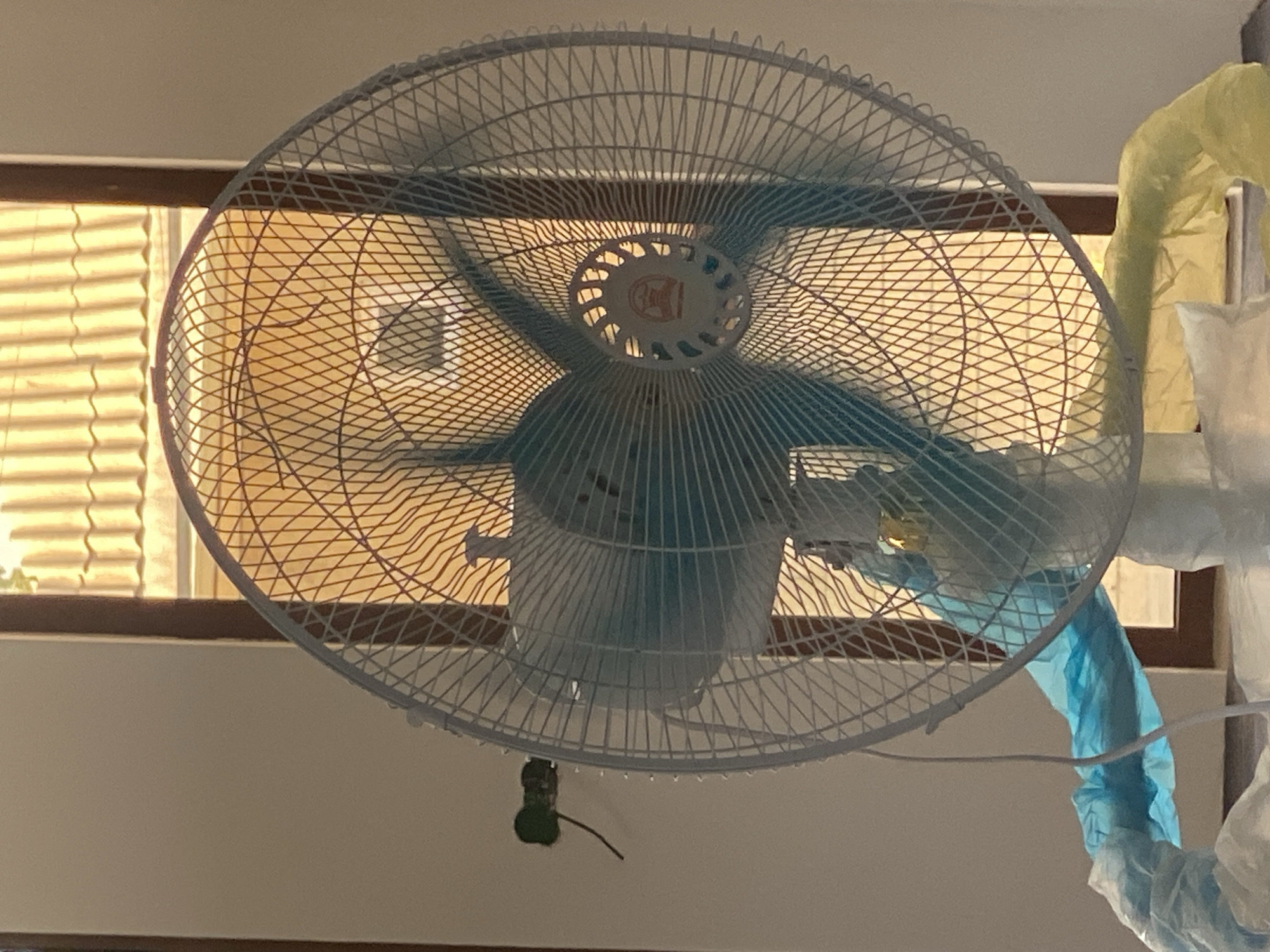 The image shows a white electric fan with blue blades, positioned in front of a window with orange blinds. The fan is partially covered with blue and yellow plastic sheeting, suggesting it is either being protected during construction or cleaning, or is being stored. A small green electrical outlet with a wire is visible on the wall to the left of the fan. The overall lighting suggests it is daytime, with sunlight filtering through the blinds. The scene appears to be indoors, likely in a residential or commercial building, but no specific location in Arbagish, Uzbekistan, can be identified from the image itself.
