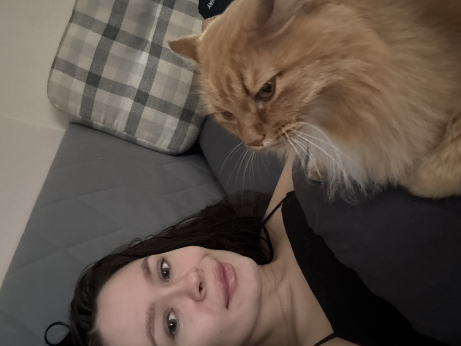 A woman with dark hair and fair skin lies on a gray sofa with a plaid pillow behind her. She wears a black top and smiles slightly at the camera. A large orange cat perches on her shoulder, its gaze directed downward with alert eyes and prominent whiskers. The scene is softly lit, capturing a moment of companionship between the woman and her feline companion.