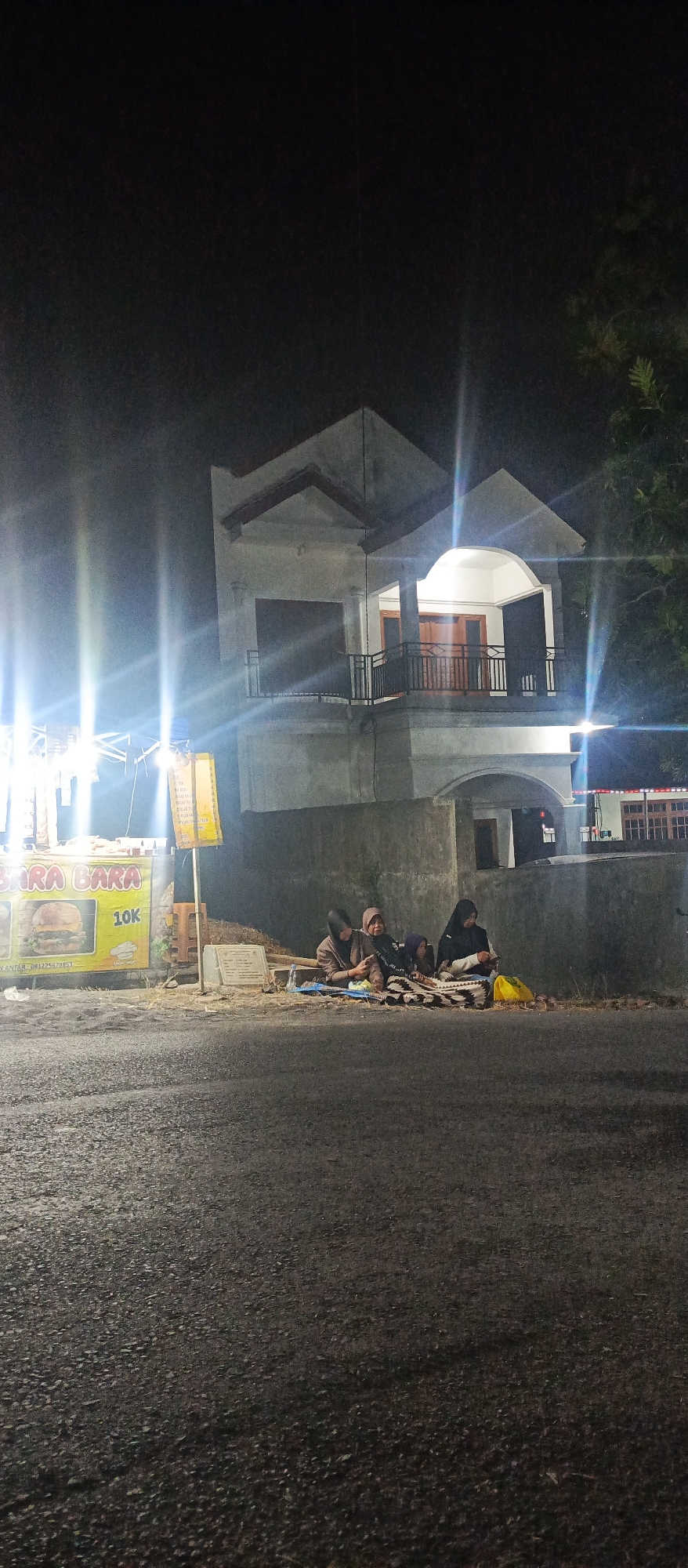 The image depicts a night scene in front of a two-story house in Sumbersekar, Indonesia. The house has a concrete facade, a balcony on the second floor with wooden doors, and is illuminated by lights. In front of the house, there is a food stall with a banner that reads "BARA BARA" and displays an image of a burger with the price "10K". To the right of the stall, four individuals are seated on the ground, facing a blanket. One individual appears to be holding a phone. A yellow plastic bag is visible to the right of the group. The ground in front of the house and stall is unpaved, and the road in the foreground is asphalt. Trees are visible on the right side of the image, and bright lights from what appear to be streetlights or a stall's lighting are casting strong beams. The overall atmosphere is that of a roadside eatery at night.