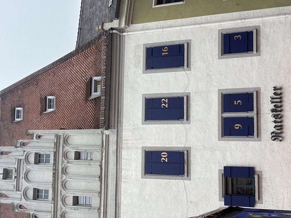 The image displays the exterior of a multi-story building facade in Meissen, Germany, under an overcast sky. The building's lower and mid-sections feature a textured white stucco finish. On the upper floor, three windows are visible. From left to right, these windows are fitted with closed dark blue shutters bearing gold numbers: "20" on the left, "22" in the center, and "16" on the right. On the floor below, three additional windows are present. The leftmost window has partially open dark blue shutters, revealing a dark interior. The central window features closed dark blue shutters, with a gold "9" on the left shutter and a gold "5" on the right shutter. The rightmost window on this level has closed dark blue shutters, with a gold "3" visible on the right shutter. Directly below the central lower window, a dark-colored sign with the word "Ratsteller" in an ornate, old German script is mounted on the facade. Above the white stucco section, an ornate white stone or stucco gable with multiple arched recesses and small, rectangular windows extends upwards. To the right of this gable, a steeply pitched roof covered in red tiles is visible, featuring several small dormer windows. A portion of an adjacent building, with a lighter-colored wall and a dark tiled roof, is visible on the far right, separated by a white vertical downspout. The scene contains no individuals or active elements.