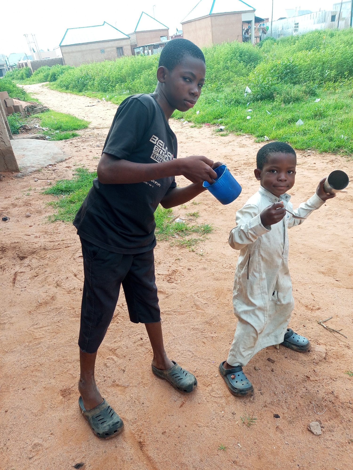 In Tempure, Nigeria, two young boys stand on a dirt path surrounded by dry grass and scattered debris. The boy on the left, older and wearing a black t-shirt and dark shorts, holds a blue plastic cup in his right hand. The younger boy, dressed in a pale, long-sleeved garment, stands to his right. He holds a silver metal cup in his right hand and appears to be interacting with something on a spoon in his left hand, possibly eating. In the background, several buildings with light-colored walls and blue-tiled roofs are visible, interspersed with overgrown green vegetation. The sky is overcast.