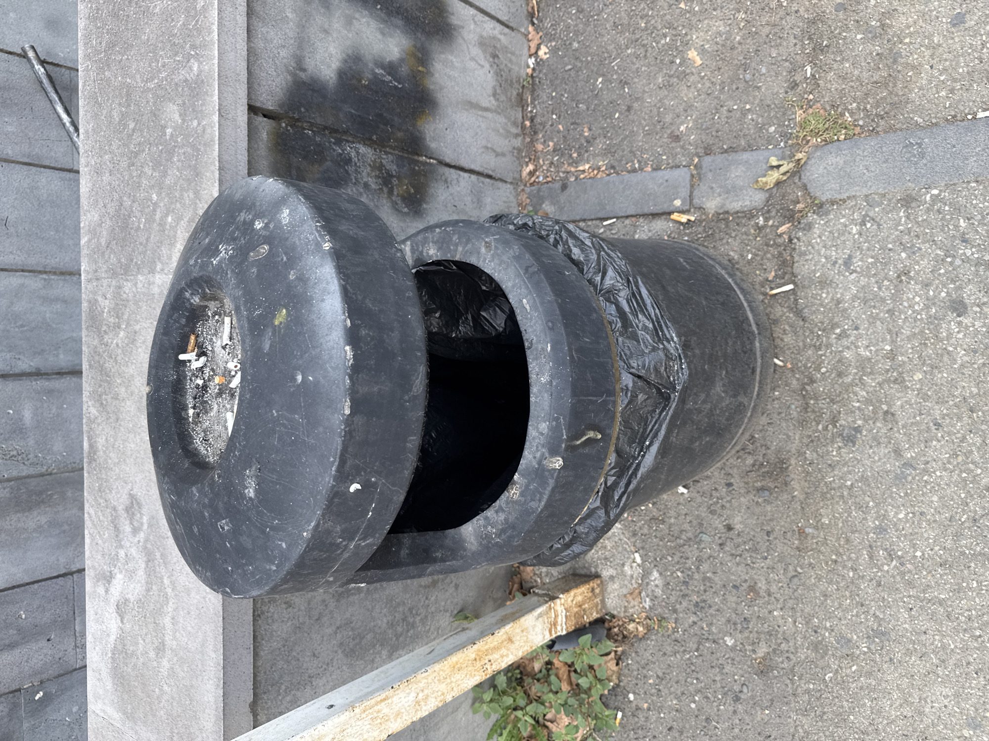 The image shows a black outdoor ashtray bin with a black plastic trash bag lining the inside. The ashtray section on top is filled with cigarette butts. The bin is located on a concrete sidewalk next to a grey stone wall. Some dry leaves and weeds are visible on the ground around the bin. The lighting suggests it is daytime. The location is identified as Tbilisi, Georgia.