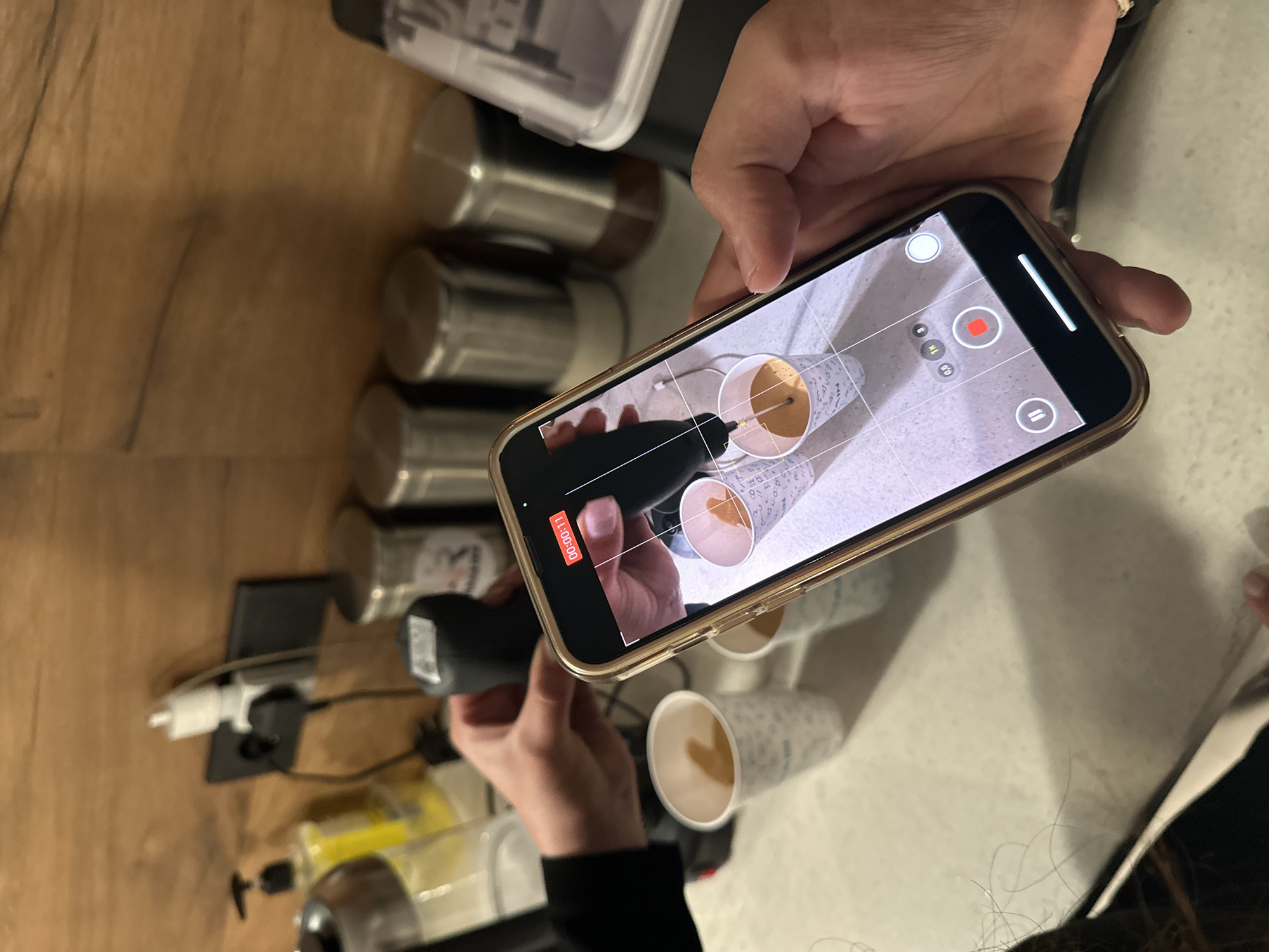 The image shows a person filming a video with an iPhone. The phone displays a recording in progress of two paper cups filled with liquid, being stirred with a handheld electric frother. The person holding the phone is also holding the frother. Several more paper cups are visible on the counter along with bottles and metal containers. The location is not identifiable as Yerevan, Armenia from the given visual information.