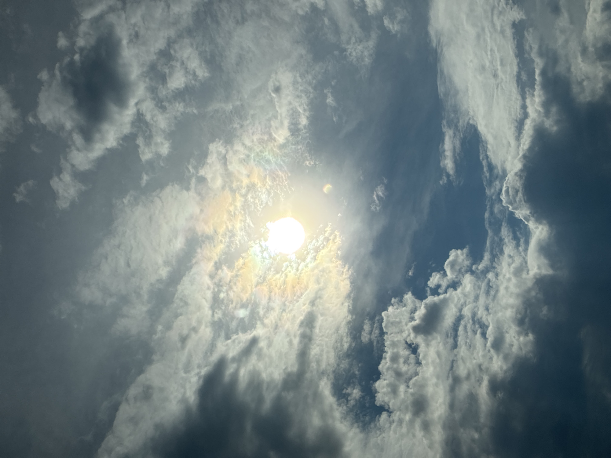 A low-angle shot captures the sun partially obscured by clouds, creating a rainbow-like effect around its bright orb. The sky is a mix of dark and light clouds, with patches of blue sky visible. The clouds are diverse, ranging from wispy streaks to dense, shadowed formations. The overall lighting suggests it is daytime, possibly late afternoon or early morning due to the angle of the sun and the dramatic cloud cover. The scene is entirely natural, with no human presence or man-made objects visible. The image is taken outdoors, looking upwards into the sky.
