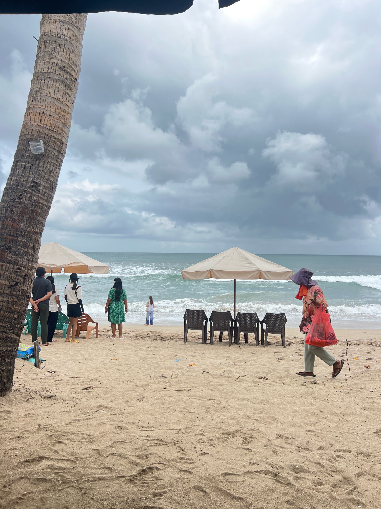 A group of people are standing on a sandy beach in Kuta, Indonesia. The sky is overcast with dark clouds. In the foreground, a large palm tree trunk is visible on the left. Several individuals are gathered near beach umbrellas and chairs. One person in a green dress is standing on the sand, looking towards the ocean. Another person wearing a wide-brimmed hat and a floral shirt is walking across the beach, carrying a red plastic bag. The ocean has moderate waves, and the beach is wet in some areas. Beach chairs are set up in front of an umbrella, and another umbrella is visible further down the beach. The sand is textured with footprints and debris.