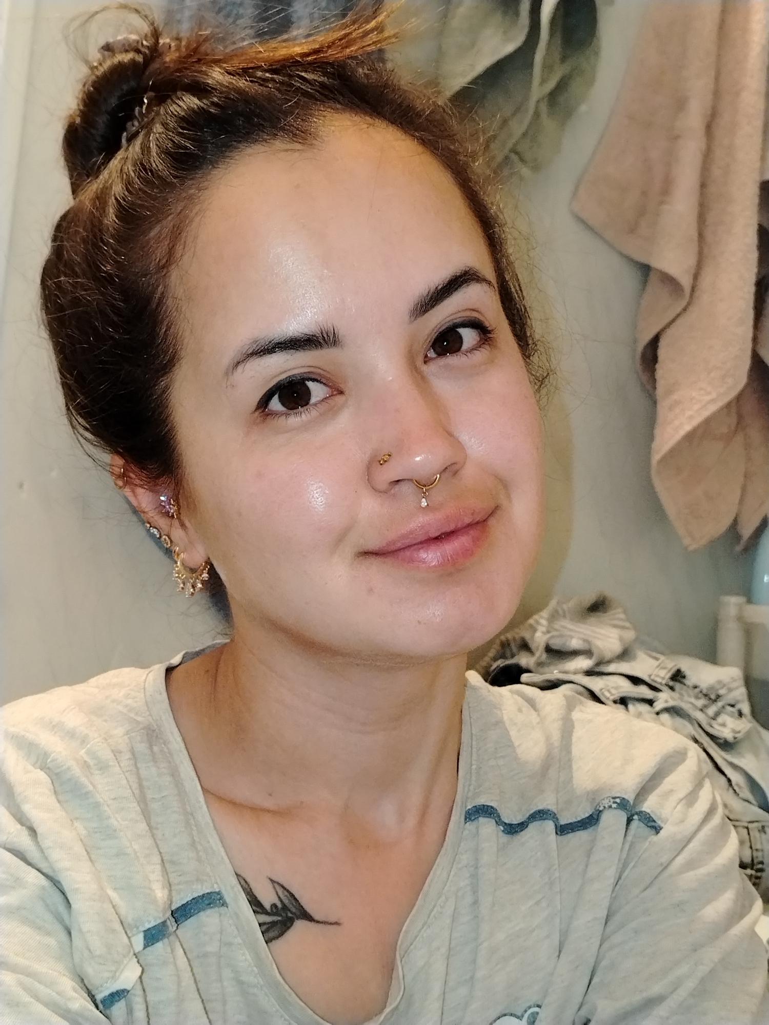 A portrait-style image captures a woman from the chest up, smiling gently at the camera in what appears to be a casual indoor setting, specified to be in Noviy, Russia, though this cannot be visually confirmed from the image. Her medium-brown hair is pulled back into a messy bun, and she has dark brown eyes and a clear complexion.
She is wearing a light-colored (possibly beige or light grey) t-shirt featuring distinctive blue wavy stitching on the shoulders. Notable accessories include multiple piercings: a golden septum ring with a small dangling clear gem, a small golden stud on her right nostril, and several earrings in her left ear, including a small purple or clear stud and a golden drop earring with small embellishments. A black ink tattoo depicting a leafy branch design is visible on her upper chest, peeking out from under her shirt collar.
The background shows a light-colored wall, with a textured, folded fabric (possibly a towel) hanging in the upper right. Below it, a pile of folded clothing, resembling denim, is visible, along with a hint of a white bottle. The lighting suggests an indoor setting, likely well-lit, perhaps by artificial light or indirect daylight. The woman's expression is calm and pleasant. There is no visible text in the image.
