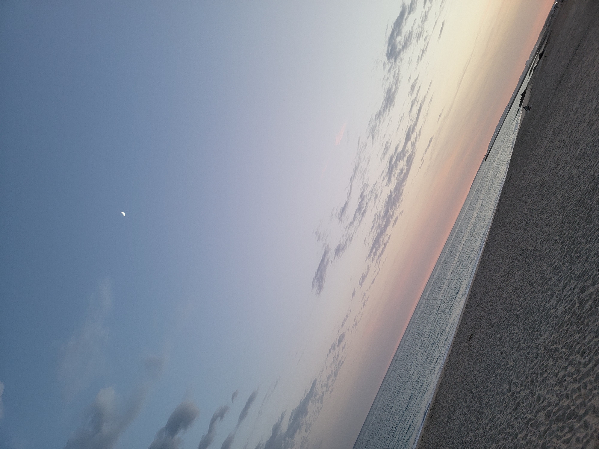 A tilted view of a beach in Quarteira, Portugal, at dusk. The sandy beach occupies the lower half of the frame, with faint footprints visible. The ocean, calm and reflecting the fading light, forms the middle ground. The sky above transitions from a pale pink near the horizon to a soft blue, with scattered wispy clouds. A crescent moon is visible in the upper portion of the sky. In the distance, along the coastline, the outlines of buildings and a pier are discernible. A few distant figures are present on the beach and near the water's edge. The overall impression is one of a serene evening by the sea.