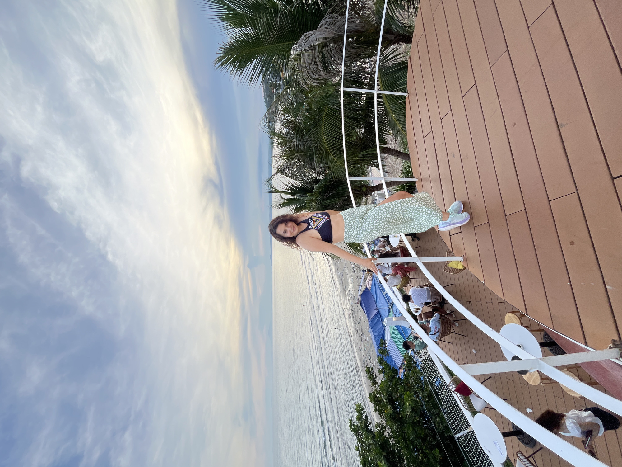 A woman in a bikini top and a long, patterned skirt stands on a wooden deck, leaning on a white railing. Behind her, the ocean stretches to the horizon under a cloudy sky. A beach is visible below the deck, with several rows of colorful tarps and umbrellas. Palm trees frame the scene on the right. Several people are seated at tables on a lower level of the deck, suggesting a restaurant or bar setting. The woman is positioned in the foreground, looking towards the camera. The overall atmosphere is relaxed and tropical. The location is likely a beachfront establishment in Mũi Né, Vietnam, known for its beaches and resorts.