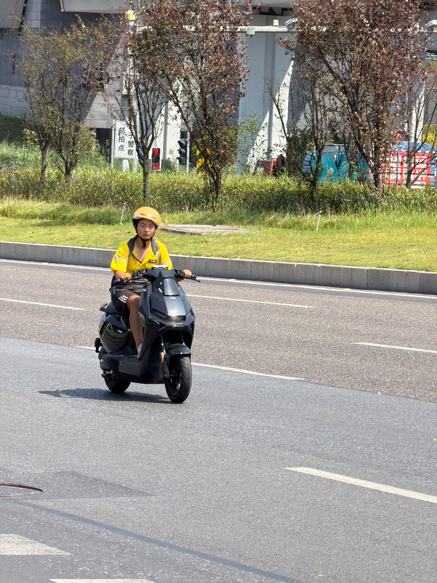 In Jiangjin, China, a person wearing a yellow polo shirt and black shorts, along with a yellow helmet, is riding a black electric scooter on an asphalt road. The scooter has a "niu" logo on the front. The person appears to be a delivery worker. The road is bordered by a concrete curb and a grassy area with trees. In the background, there are buildings and traffic signals, including one that is red. The sky is clear and the sun is shining, casting shadows.