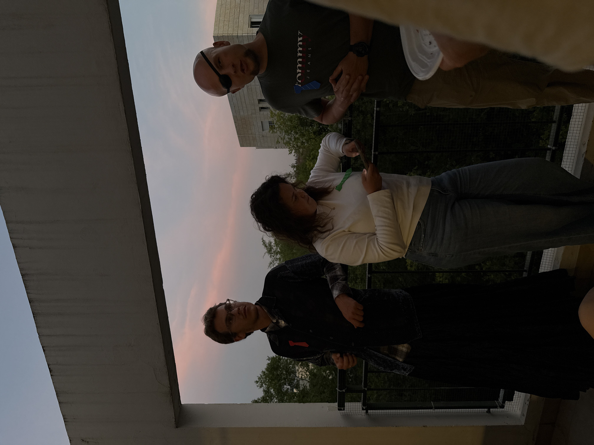 The image shows three people on a balcony overlooking a cityscape at dusk. The location is likely Modiin-Maccabim-Reut, Israel, based on the urban architecture and environment.
The person closest to the camera is wearing glasses, a plaid shirt, and a dark jacket. He is standing near the balcony railing. Next to him is a person wearing a long-sleeved white shirt and jeans. They appear to be looking at something in their hands, potentially a phone. At the top of the frame, a man with an eye patch is wearing a green Tommy Jeans shirt and khaki pants. He appears to be talking or posing for the camera.
The environment includes a concrete balcony railing and a view of buildings and trees against a darkening sky with a pinkish hue. The lighting suggests it is either late afternoon or early evening. A white bowl or container is being held by someone in the top of the frame, outside of the area of focus.