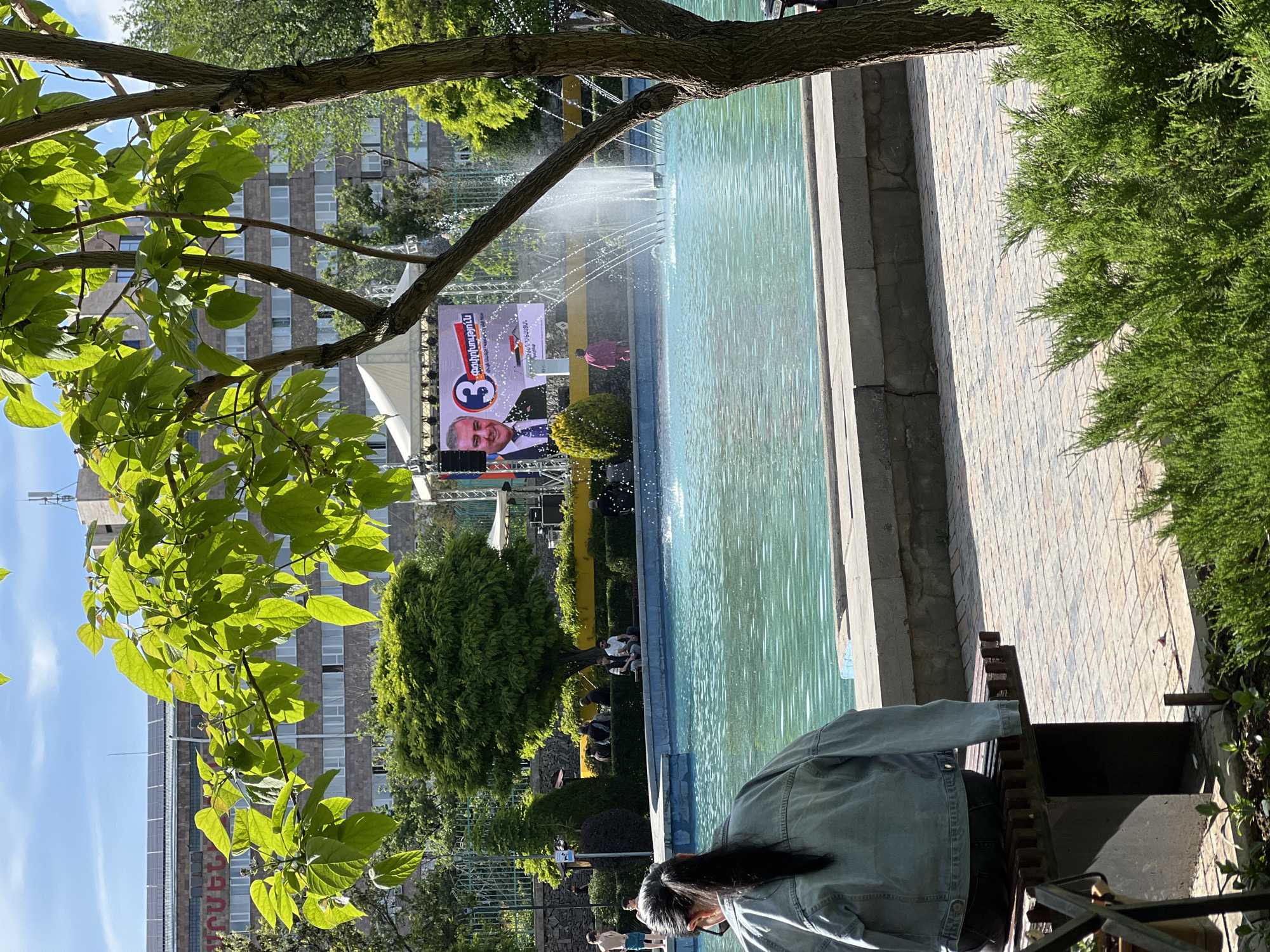 The image depicts a scene in Yerevan, Armenia. In the foreground, a person with long dark hair and wearing a blue denim jacket is sitting on a park bench, seemingly looking down at something in their hands. Beyond the bench is a large rectangular fountain filled with water and spraying a water feature into the air. Behind the fountain is a stage set up with a large screen showing a man in a suit with a campaign advertisement. There are people seated in front of the stage. The environment includes trees with lush green leaves framing the scene. A multi-story building is visible in the background. The ground is paved with bricks near the fountain.