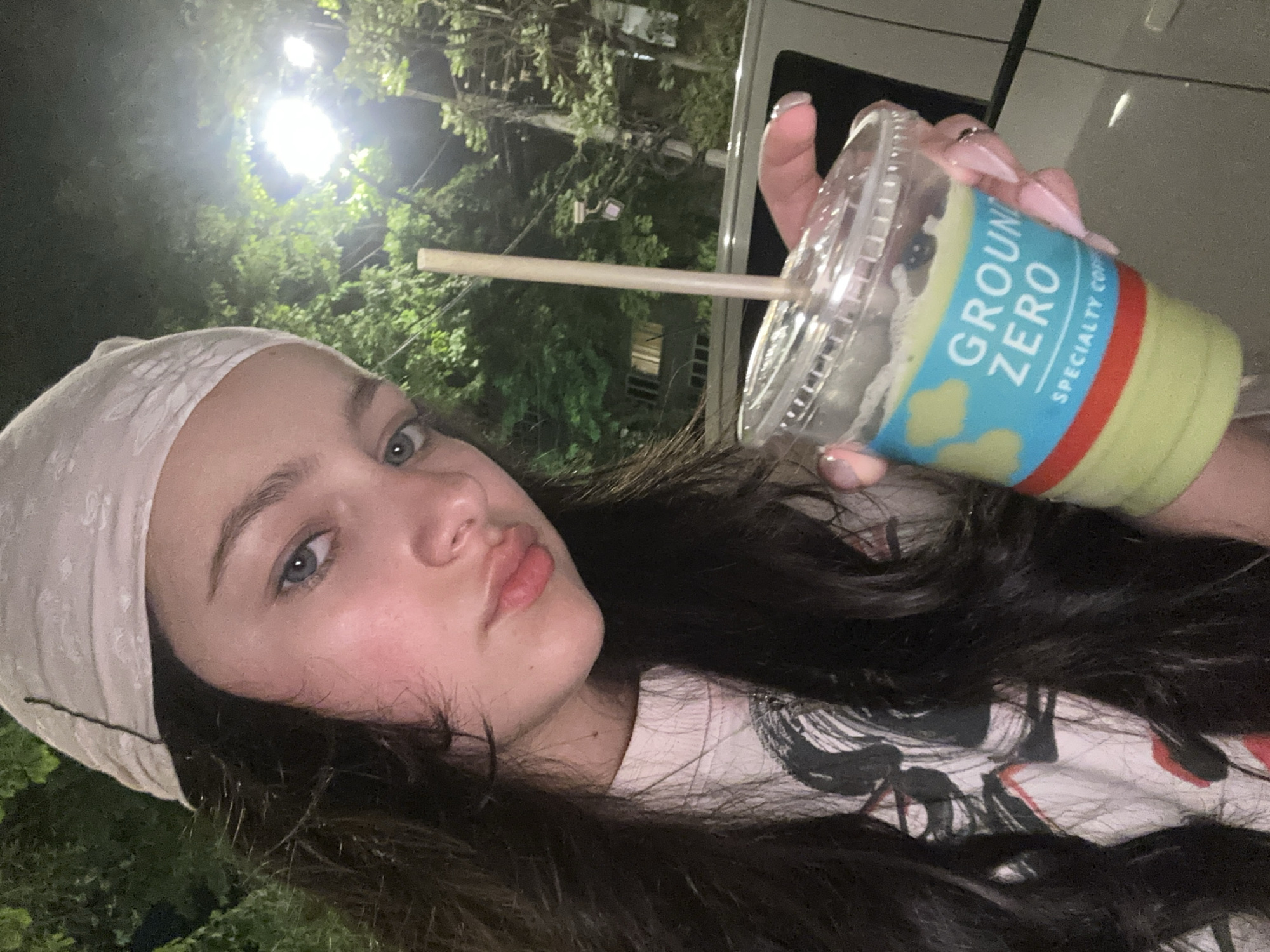 A young woman poses for a selfie while holding a partially consumed green-colored beverage from "Ground Zero Speciality Coffee", likely in Yerevan, Armenia, based on the establishment's location. She is wearing a white patterned headscarf and has dark hair. The woman is wearing a t-shirt, partially obscured by her hair, that has a black and white design on a white background. The background shows an outdoor setting with trees, streetlights, and what appears to be a light-colored vehicle.