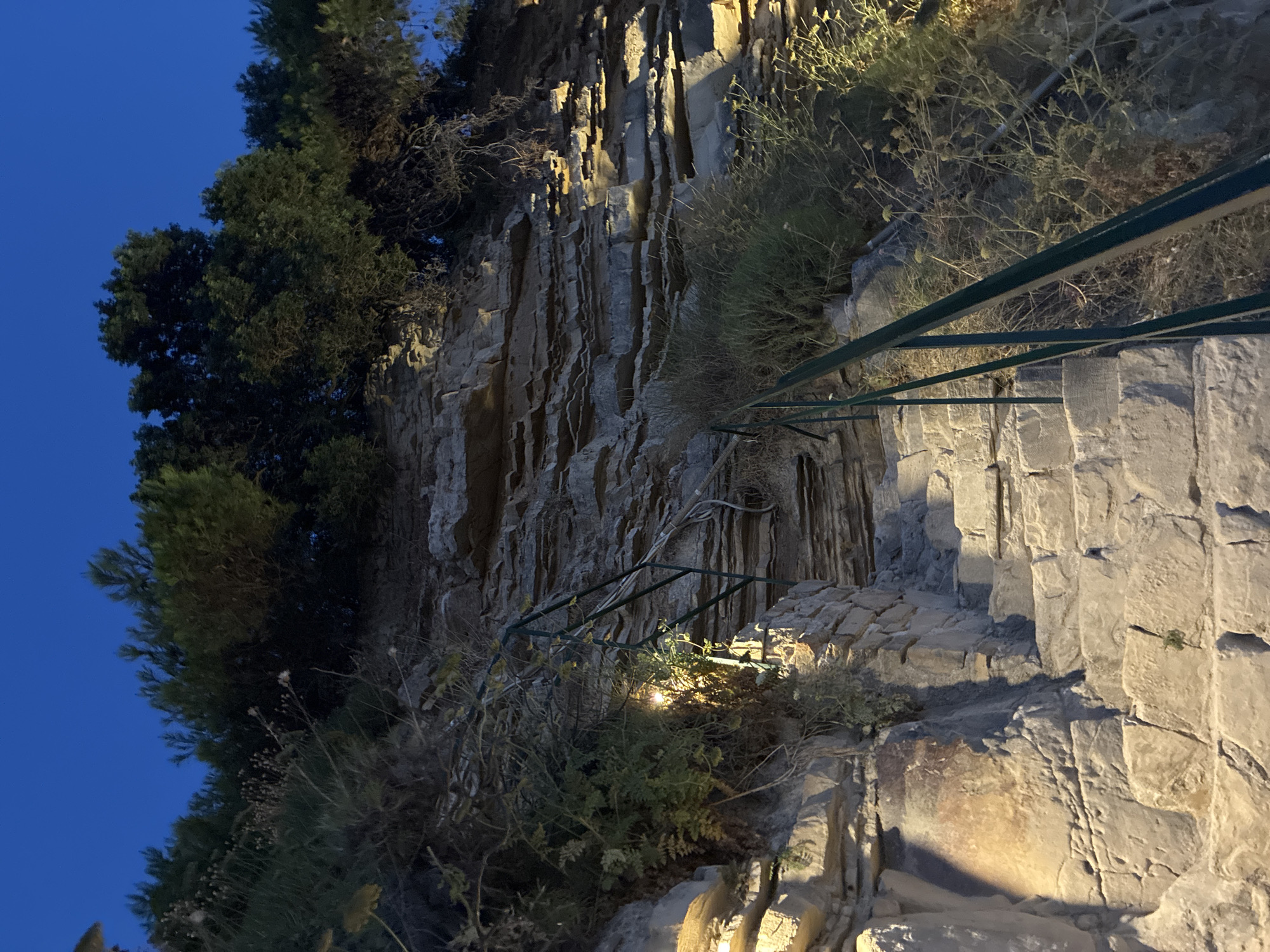 The image shows a stone staircase leading up a rocky embankment at dusk. The embankment is covered with sparse vegetation and has visible layers of sedimentary rock. A green metal railing runs alongside the stairs. The sky above is a deep blue, indicating twilight. There are no people or animals visible in the scene. The lighting suggests that artificial lights are illuminating the staircase and parts of the embankment. The location is in the Municipal Unit of Laganas, Greece, likely a coastal area given the rocky terrain and vegetation.