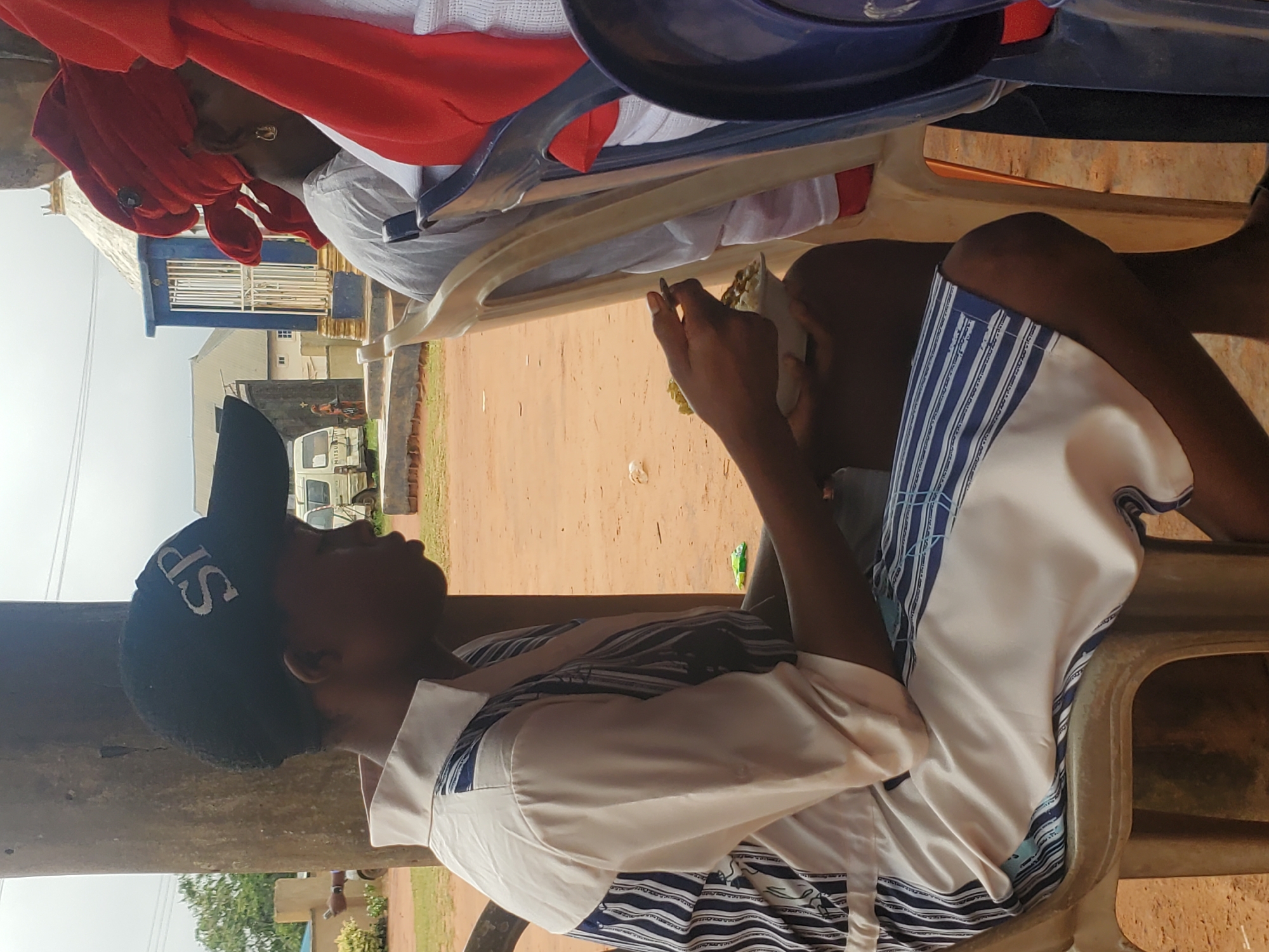 The image shows a side profile of a young person wearing a black baseball cap with white lettering that appears to say "SP" and a white shirt with blue vertical stripes. They are seated in a beige plastic chair, holding a small white bowl of food and a utensil, and appear to be eating. To the right of this person, another individual is visible from the side, wearing a red head covering and a white garment. The background suggests an outdoor setting with a dirt ground, a wooden pole, and some buildings in the distance. A white van is also visible parked in the background. The scene appears to be in Nsugbe, Nigeria, likely a public or communal gathering.