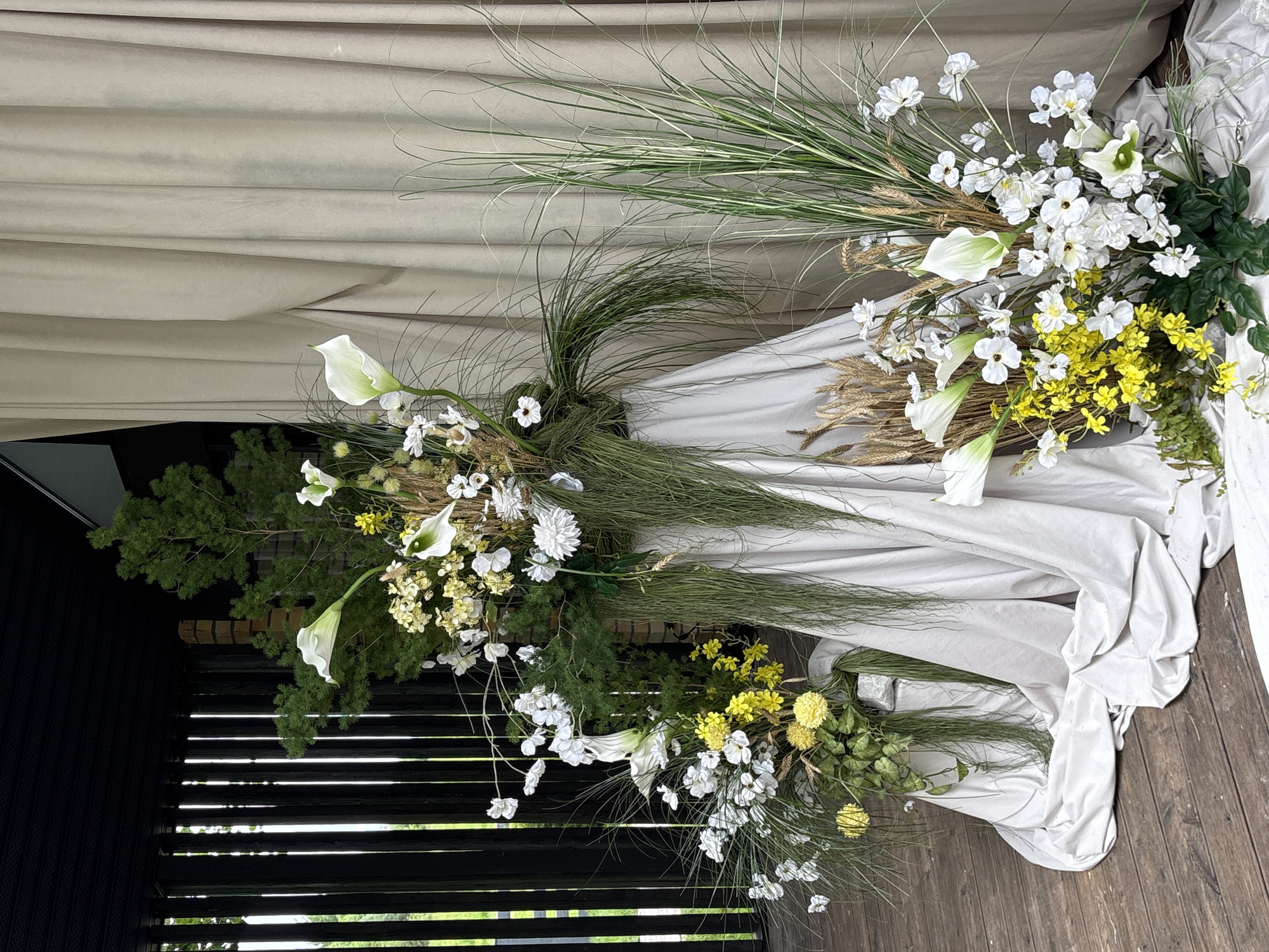 The image depicts an elaborate floral display set up in an outdoor or semi-outdoor location within the city of Perm, Russia. The scene features multiple arrangements composed of white, yellow, and green elements, set against a backdrop of draped fabric and dark wooden slats on a wooden deck.
On the left side, a dark, slatted wooden wall or fence extends vertically, partially revealing glimpses of outdoor greenery. Above, a dark, flat overhead structure is visible. A medium-sized floral arrangement featuring white and yellow blossoms, along with various green foliage, is situated in front of this slatted wall, resting on a light-colored fabric drape that pools onto the wooden deck.
Centrally, a tall and prominent floral arrangement stands, its base concealed by a draped, light beige fabric that forms soft folds on the deck. This arrangement incorporates tall, dark green coniferous branches, white calla lilies, clusters of small white and yellow flowers, dried wheat stalks, and long, cascading strands of dark green foliage.
To the right, a large, light beige fabric drape serves as a backdrop, spanning the height of the image with soft, vertical folds. In front of this drape, a tall cluster of variegated green and white striped grasses stands. Towards the lower right, another floral arrangement is positioned on the draped fabric. This lower arrangement includes white calla lilies, numerous small white flowers, bright yellow blossoms, and green leafy plants at its base, interspersed with dried wheat stalks.
The ground across the entire visible area is a dark brown wooden deck, with planks laid in a horizontal and slightly diagonal pattern. The overall presentation suggests a decorative setup for an event or display.