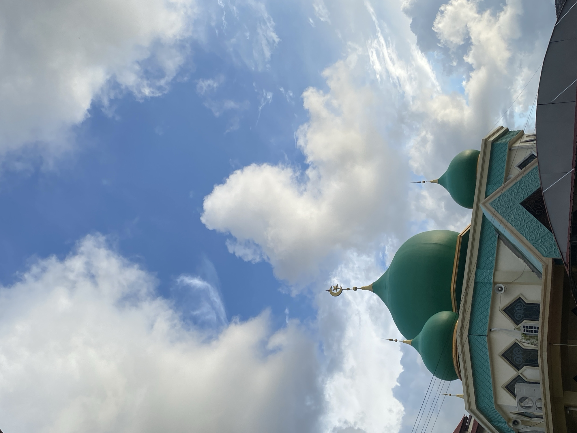 The image shows a mosque in Banda Aceh, Indonesia, with a bright blue sky and white clouds. The mosque has several green domes, the largest of which is adorned with a golden crescent moon and star. The building's architecture features intricate geometric patterns in turquoise and white. In the foreground, a section of the mosque's roof is visible, covered by a gray awning. The overall scene conveys a sense of tranquility under a partly cloudy sky.