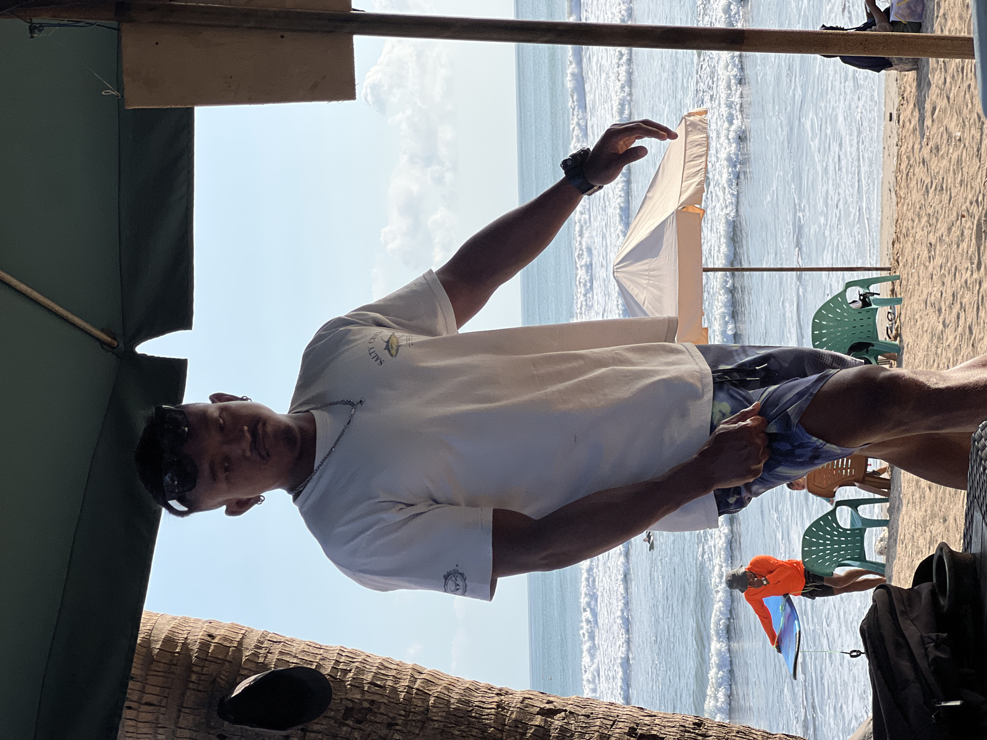 The image depicts a beach scene in Kuta, Indonesia, under a clear blue sky with moderate waves. The foreground is dominated by a person wearing a white t-shirt and patterned shorts, standing partially obscured by a palm tree and an overhead canopy. This individual has sunglasses perched on their head and a watch on their left wrist. In the middle ground, another person in an orange long-sleeved shirt and dark shorts is standing near the water's edge, holding a blue surfboard. Several beach chairs are visible, some empty and some occupied by other beachgoers in the background. An umbrella provides shade on the sand. The ocean stretches out to the horizon, with small waves breaking on the shore. The overall atmosphere suggests a relaxed, tropical beach setting.