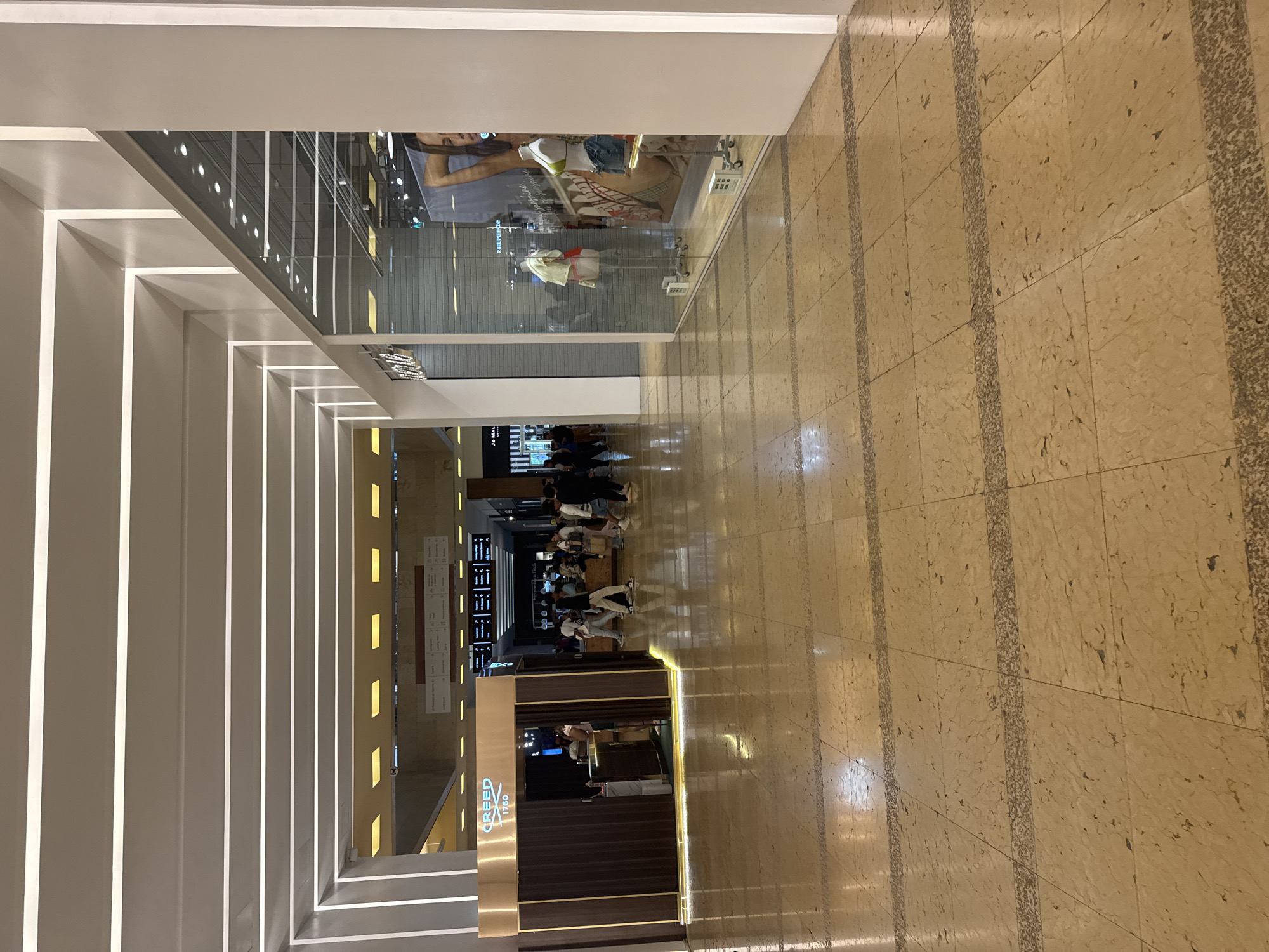 The image depicts an indoor space, likely a shopping mall, in the city of Puebla, Mexico. The camera angle is looking down a corridor, flanked by retail spaces and illuminated by evenly spaced linear light fixtures on the ceiling. The flooring consists of beige tiles with darker, textured lines creating a grid pattern.
On the left is a storefront with a display window showing a mannequin and advertisements. Further down the corridor, there are people gathered, possibly in a food court area, with tables and chairs visible. A shop named "CREED 1760" is also present, identifiable by its signage.
The scene appears to be moderately busy, with people engaging in various activities such as shopping, eating, or simply passing through. The overall ambiance is brightly lit and seems well-maintained.