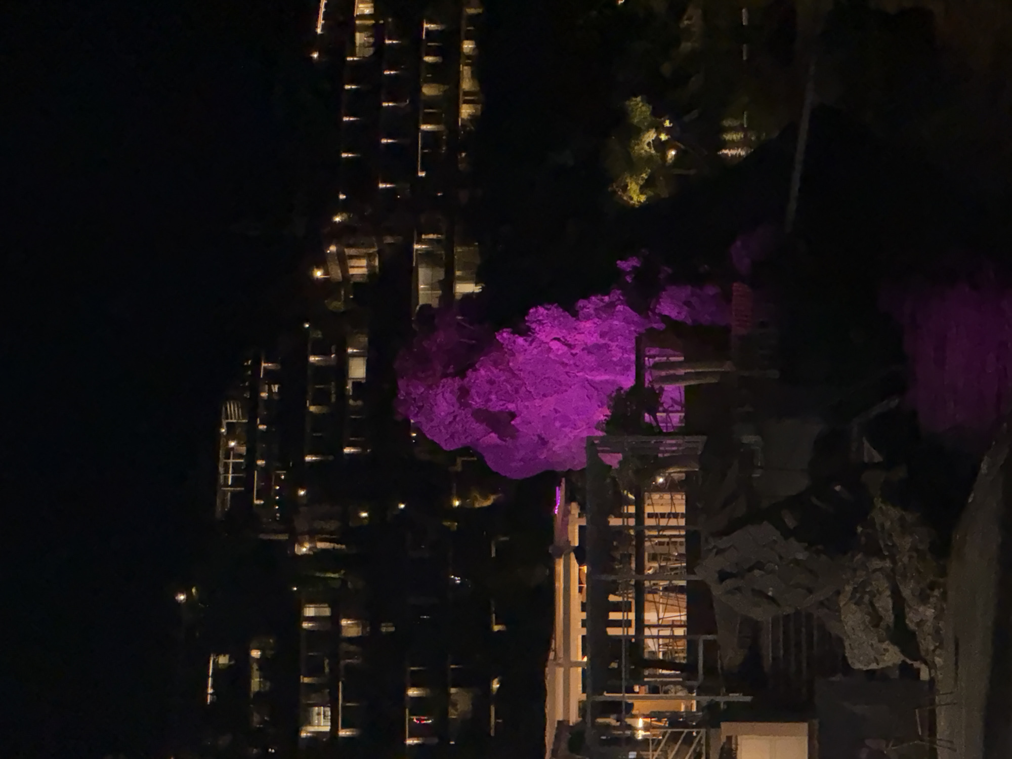 The image depicts a nighttime scene at a coastal resort, likely in Budva, Montenegro. In the foreground, large, dark rocks are visible, with some appearing wet and reflecting artificial light. A structure, possibly a restaurant or lounge area with outdoor seating and railings, is situated above the rocks. One prominent rock formation in the mid-ground is dramatically illuminated with bright pink lighting. Behind this, a hillside is covered with multi-story buildings, appearing to be hotels or apartments, with numerous windows lit from within, suggesting occupancy. The overall impression is one of a well-lit resort area at night, with the illuminated rock serving as a focal point. The sea is visible in the background, reflecting the lights of the resort and the pink-lit rock.