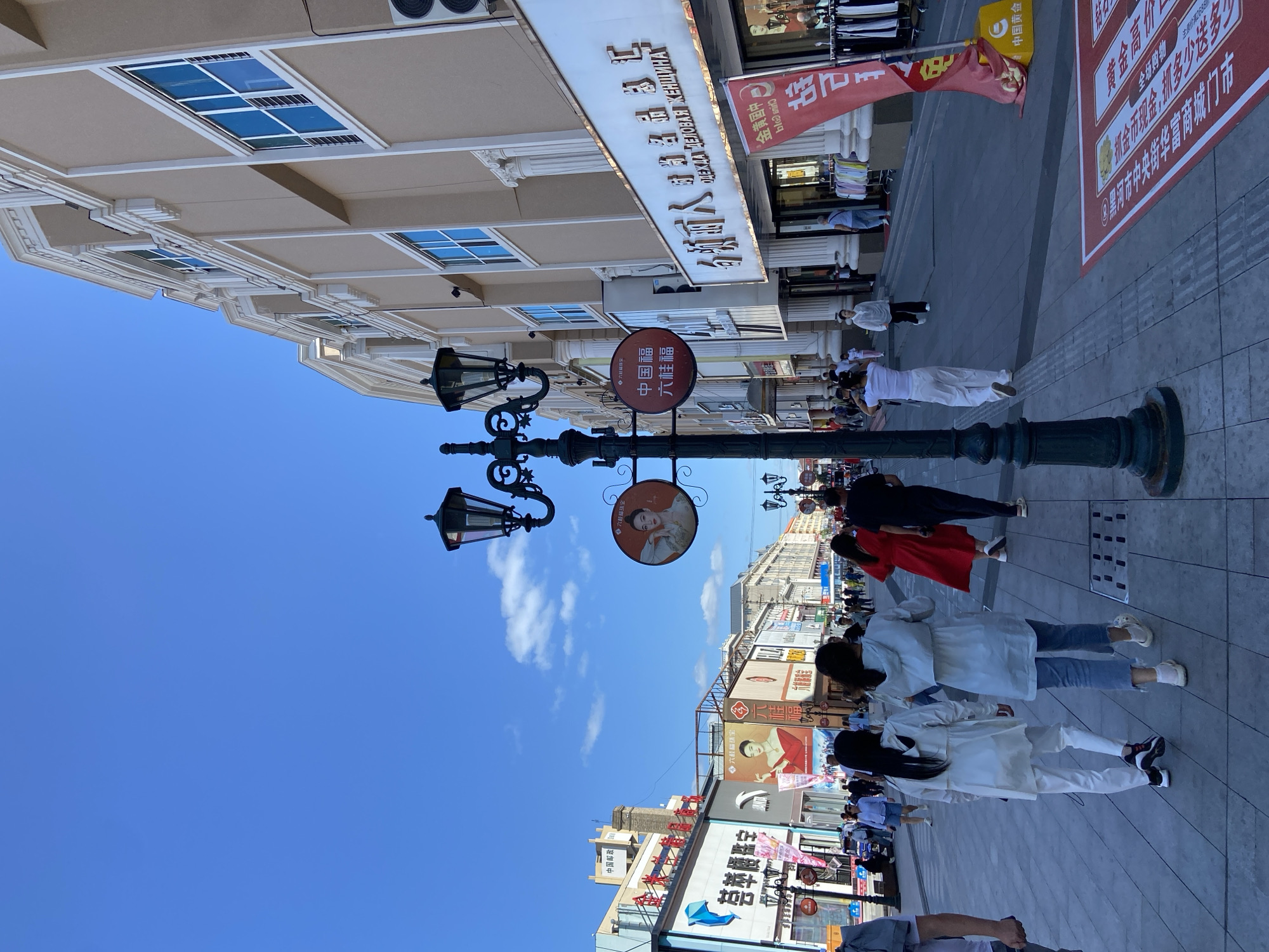 A bright, sunny day features a bustling pedestrian street lined with shops in Xing'an Subdistrict, China. The street is paved with gray tiles and divided by a yellow tactile paving strip. A tall, dark green lamppost with multiple light fixtures stands prominently in the foreground, adorned with two circular signs, one depicting a woman in traditional attire and another with Chinese characters.
On the right side of the street, a large, light beige building with classical architectural elements, including columns and ornate trim, houses shops. The windows of this building are reflective blue. A red banner with Chinese characters is displayed in front of one of the shops. Clothing racks with garments are visible outside another store.
On the left side of the street, various shops display colorful signage and advertisements, including a large billboard featuring a woman in a red dress. People are walking in both directions along the street. Among them, two women in white outfits are walking away from the camera, and a woman in a red dress is walking towards the right. Several other individuals, dressed in casual attire, are also seen strolling or browsing. The sky is a clear, vibrant blue with a few scattered white clouds.