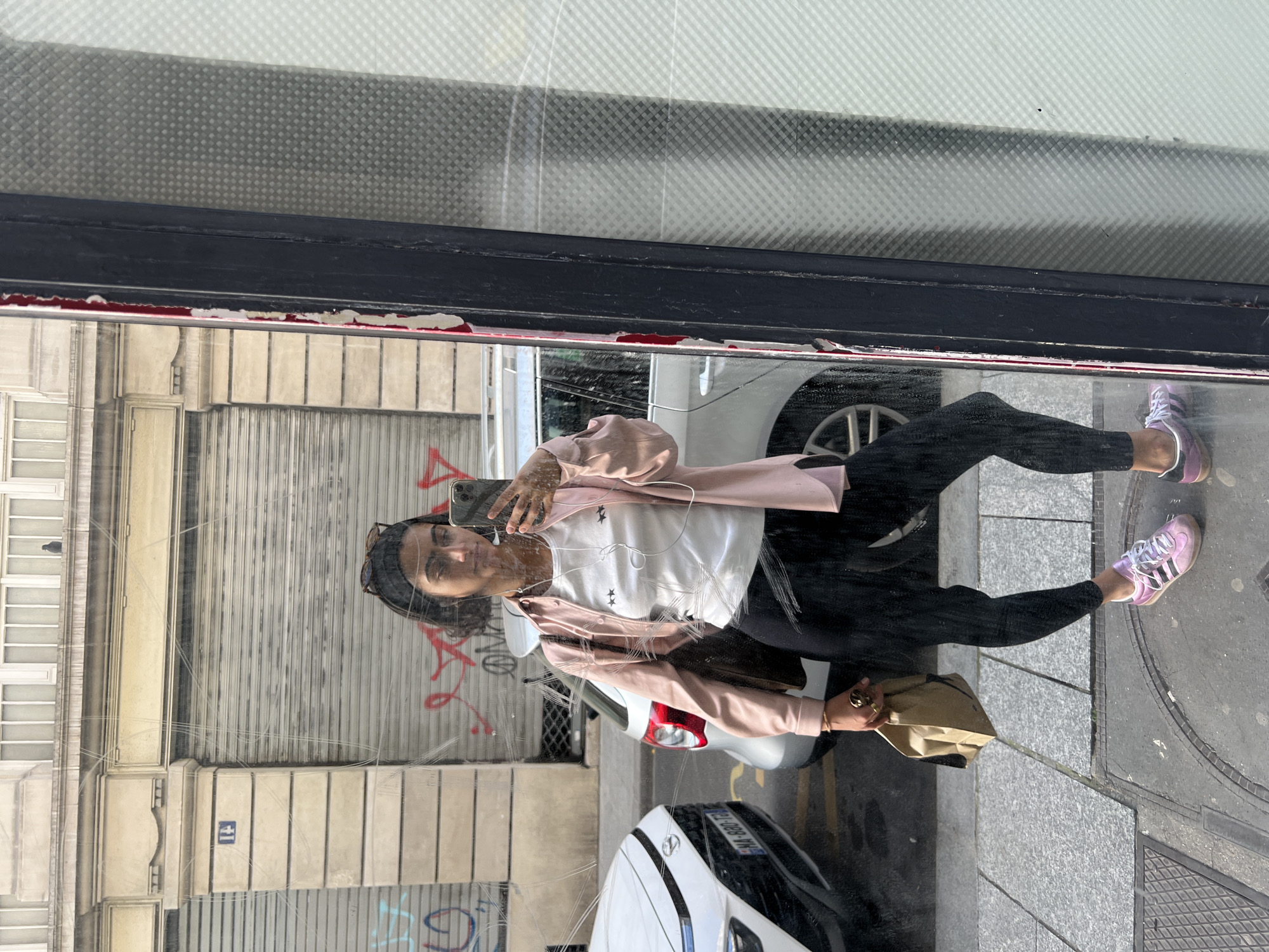 The image shows a woman taking a selfie in the reflection of a shop window in Paris, France. The reflection reveals a street scene with buildings, graffiti, a parked car, and a closed metal shutter. The woman is wearing black leggings, a white t-shirt with star graphics, a pale pink jacket, and pink Adidas sneakers. She is holding a phone in one hand and a brown paper bag in the other. The shop window has a black frame, and some of the frame has chipped off.