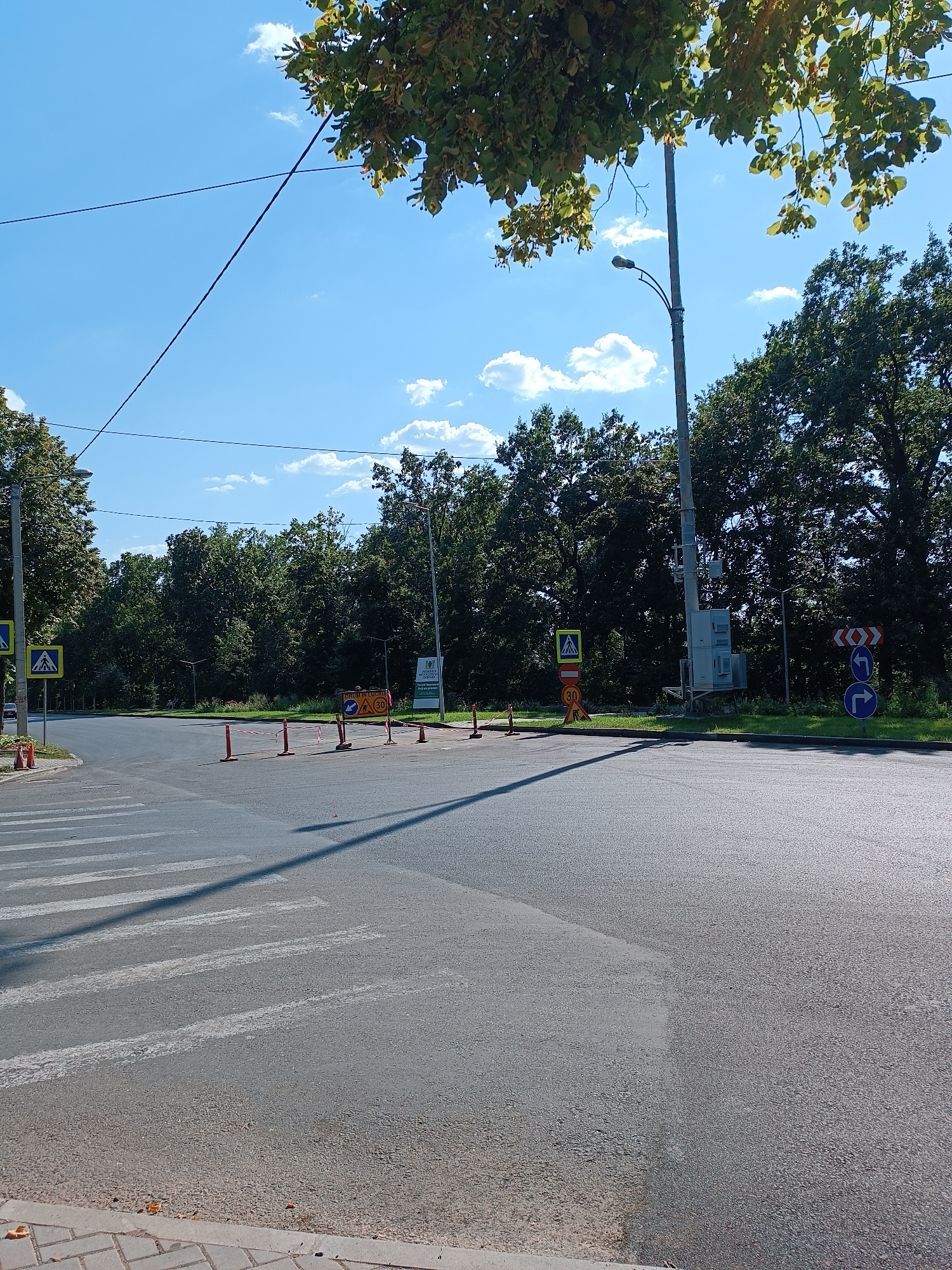 The image depicts an intersection in Chișinău, Moldova, with a pedestrian crossing in the foreground and several road signs. The sky is blue with scattered clouds, and trees line the background. There are no people or vehicles visible in the scene, suggesting a quiet moment. The road appears to be asphalt, with white lines marking the pedestrian crossing. Orange cones and tape are present, indicating some form of road work or temporary obstruction. A lamppost stands on the right side of the image. Several traffic signs are visible, including a pedestrian crossing sign, a speed limit sign of 30, and directional signs indicating turns. A large white sign with text and an emblem is also present on the right. The overall impression is of a well-maintained urban environment on a clear day.