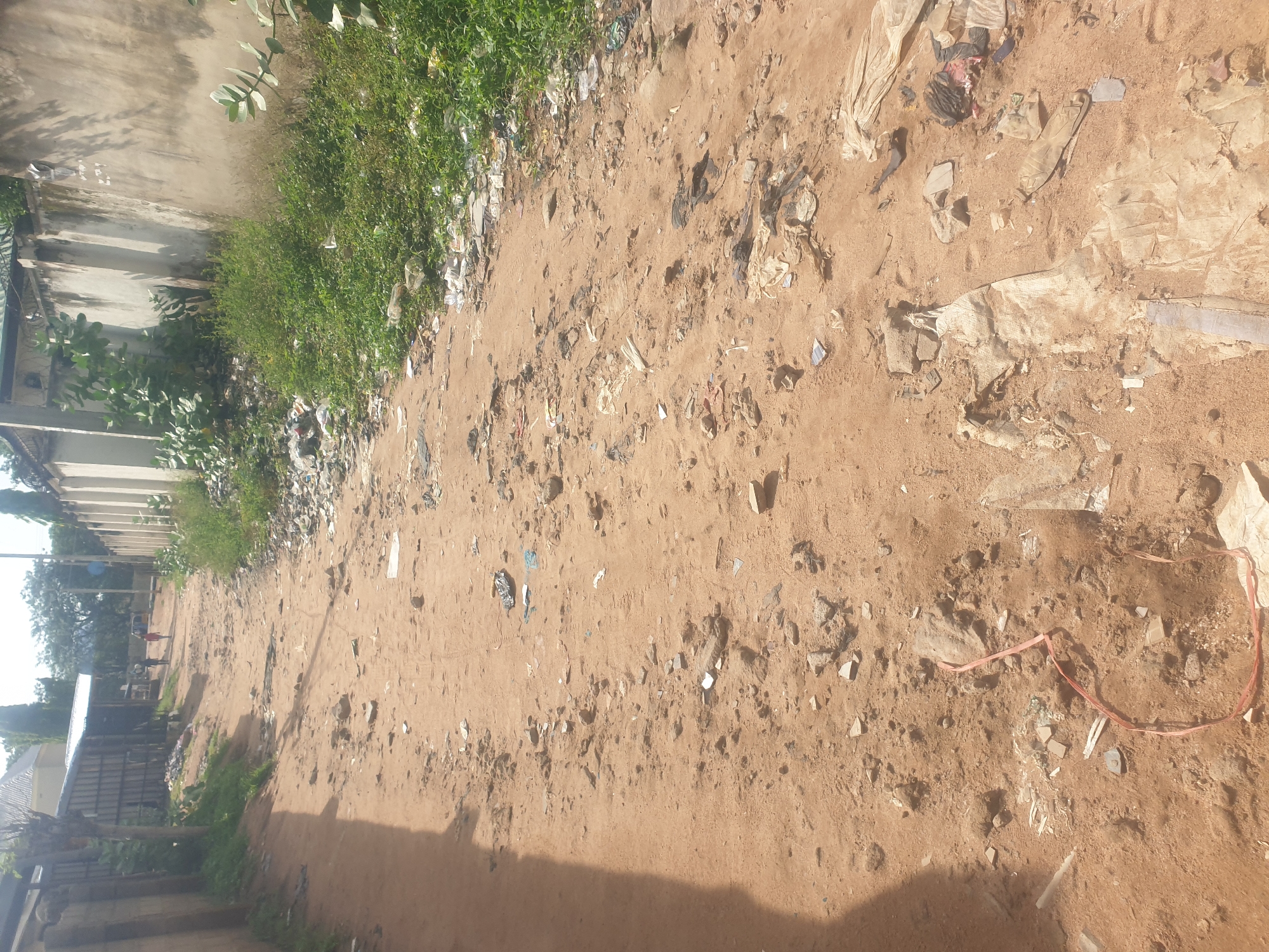 The image depicts a dirt road in Jauro Yinu, Nigeria, littered with trash. The road is unpaved and sandy, with various types of debris scattered across its surface. This debris includes plastic bags, bottles, torn fabric, and other discarded materials. The sides of the road are lined with overgrown vegetation and a concrete wall on the right. In the background, there are several people walking on the road, and a few structures, including buildings and fences, are visible. The scene appears to be an outdoor, public area within a residential or semi-urban environment, characterized by a lack of proper waste management.