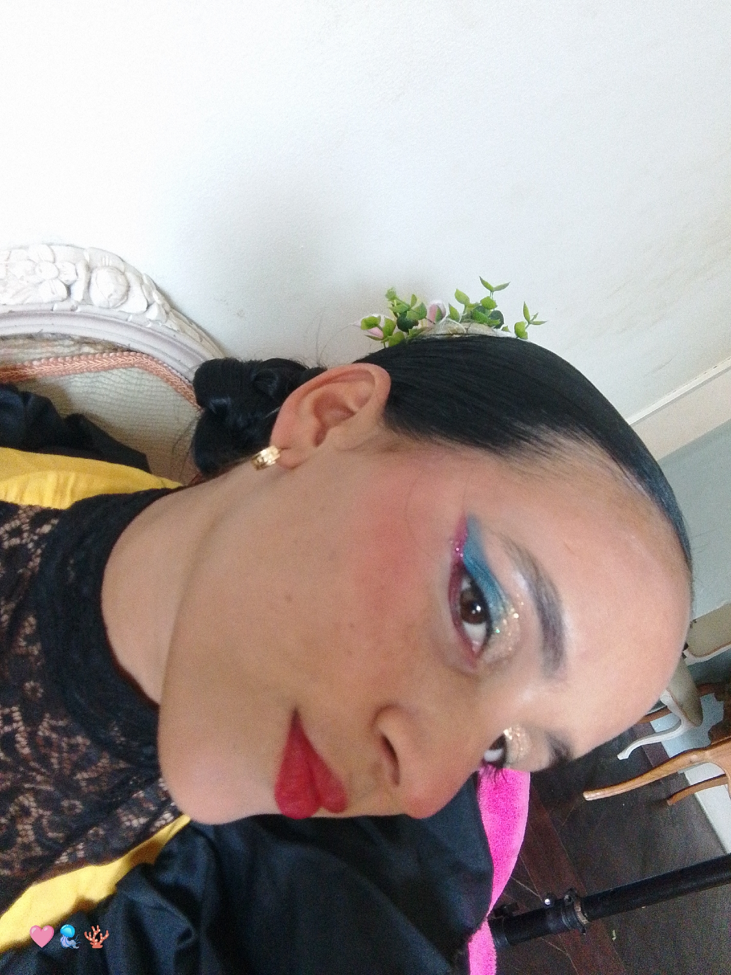 A close-up, tilted shot features a woman with fair skin, red lipstick, and elaborate eye makeup in blue, pink, and glitter, her dark hair pulled back with small flowers adorning the top of her head. She wears a black lace top with yellow accents and a black shawl. Part of a white ornate chair is visible on the left, and a dark-toned wooden floor with light-colored chairs is seen on the right. A pink object and a black metal pole are in the lower center. The setting is unspecified and there is no information to relate this image to Pedro González, Venezuela.