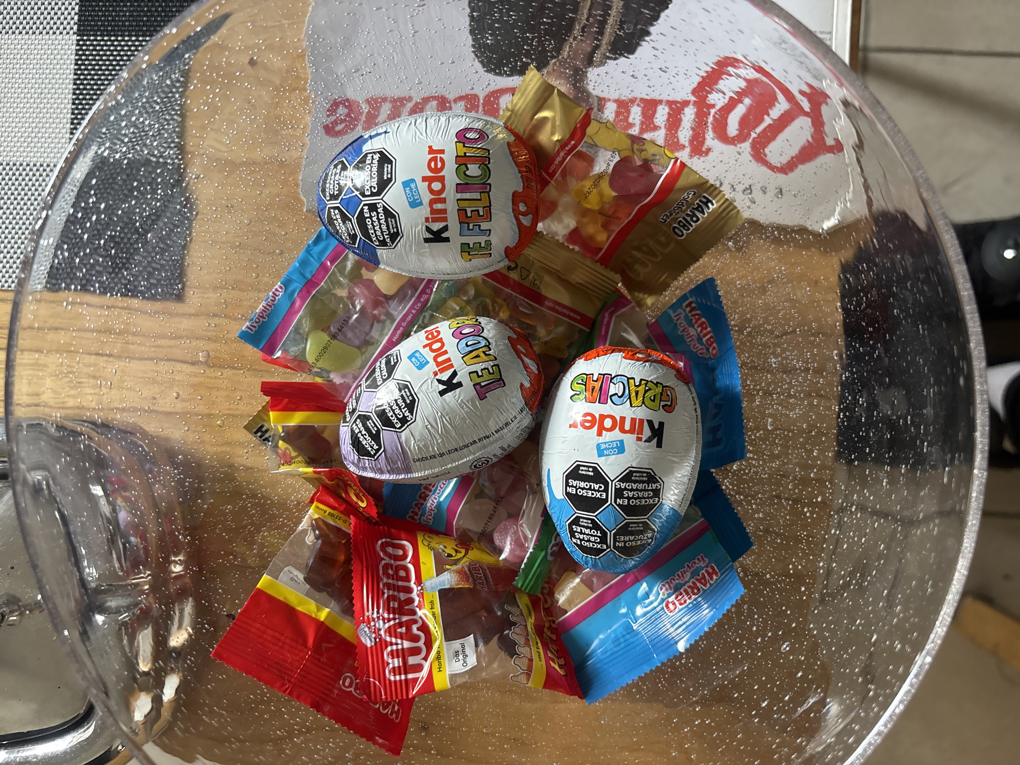 A close-up, high-angle shot shows the interior of a clear glass bowl filled with individually wrapped candies. Prominently displayed are three Kinder brand chocolate eggs with soccer ball designs on their foil wrappers. The wrappers are printed with the phrases "TE FELICITO," "TE ADORO," and "GRACIAS" in Spanish. Also present are an assortment of Haribo gummy candies, including "Tropifrutti" and "Goldbären" varieties. The bowl appears slightly wet or has condensation on the inside surface. The background is partially visible and includes a checkered surface and hints of other objects, suggesting an indoor setting.