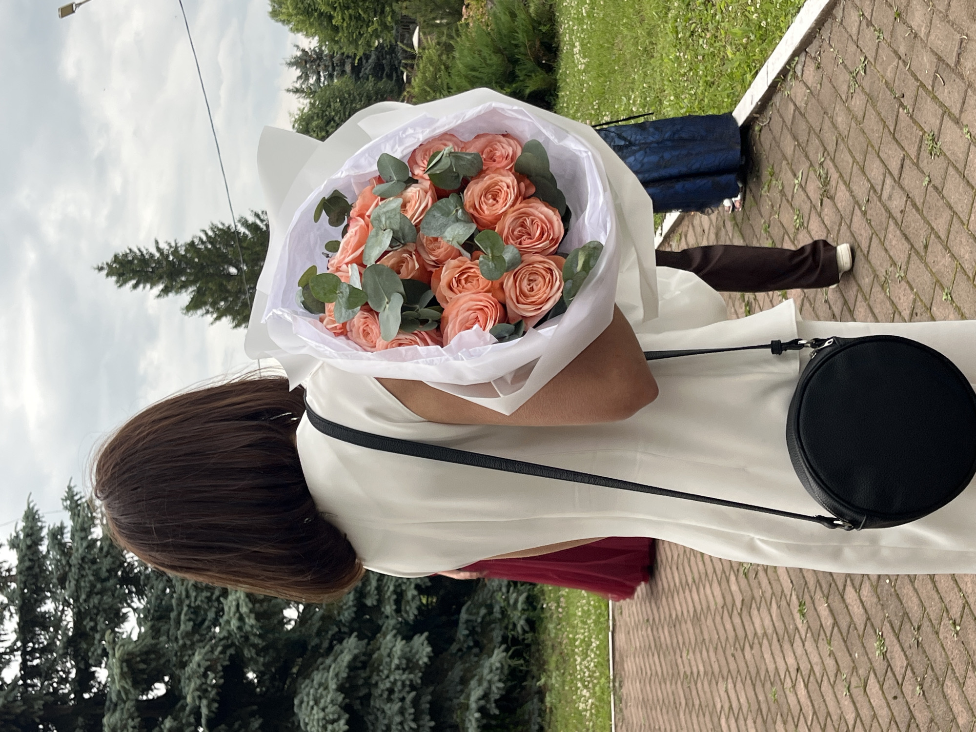 The image shows a person from the back, holding a bouquet of coral roses and eucalyptus. The bouquet is wrapped in white paper. The person is wearing a white dress and carrying a black round purse. In the background, there are evergreen trees, a brick pathway, and grass. Another person wearing a dark blue dress is visible in the background. The weather appears to be overcast. The image is a vertical shot.