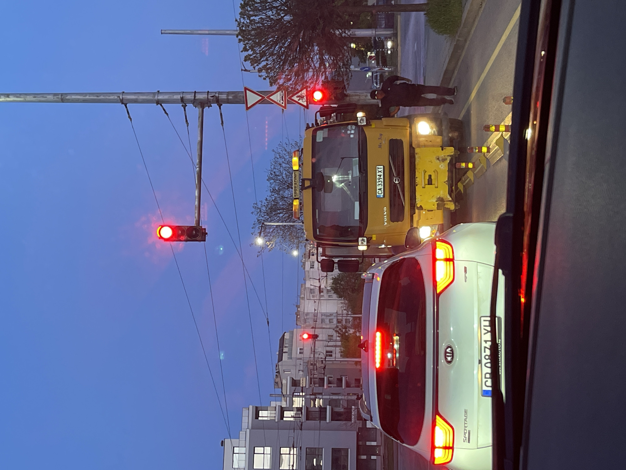 The image shows a street scene in Sofia, Bulgaria at dusk. A white Kia Sportage is stopped in traffic behind a yellow Volvo construction truck. The license plate of the Kia is CB 0871 YH. The truck has a license plate that reads CA 3394 XT. The truck is positioned near orange and white traffic cones, and a man in dark clothing is standing next to the truck. Overhead traffic lights are visible and are red. The background features apartment buildings and power lines. The photo appears to have been taken from inside another vehicle.