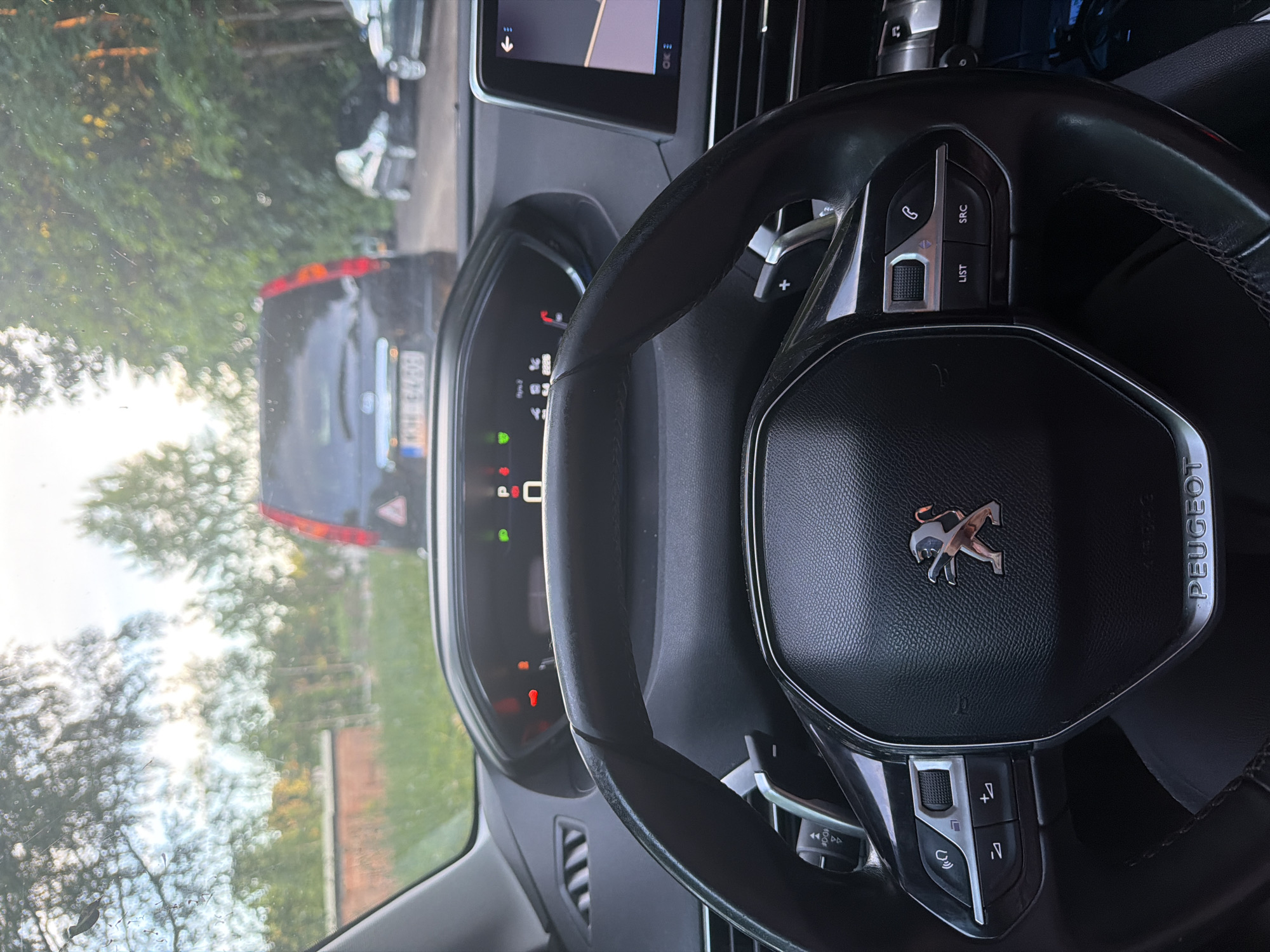 The image is a first-person perspective from inside a Peugeot car, looking out through the windshield. The car is stationary, likely in a traffic jam, as indicated by the cars visible in front. The scene outside is a rural or suburban setting with trees and a brick building. The time of day appears to be late afternoon or early evening, given the lighting conditions. The dashboard of the car is clearly visible, showing various controls and a digital display. The steering wheel is also prominent, with the Peugeot logo in the center. There are no people visible in the image. The location is not discernible from the image itself.