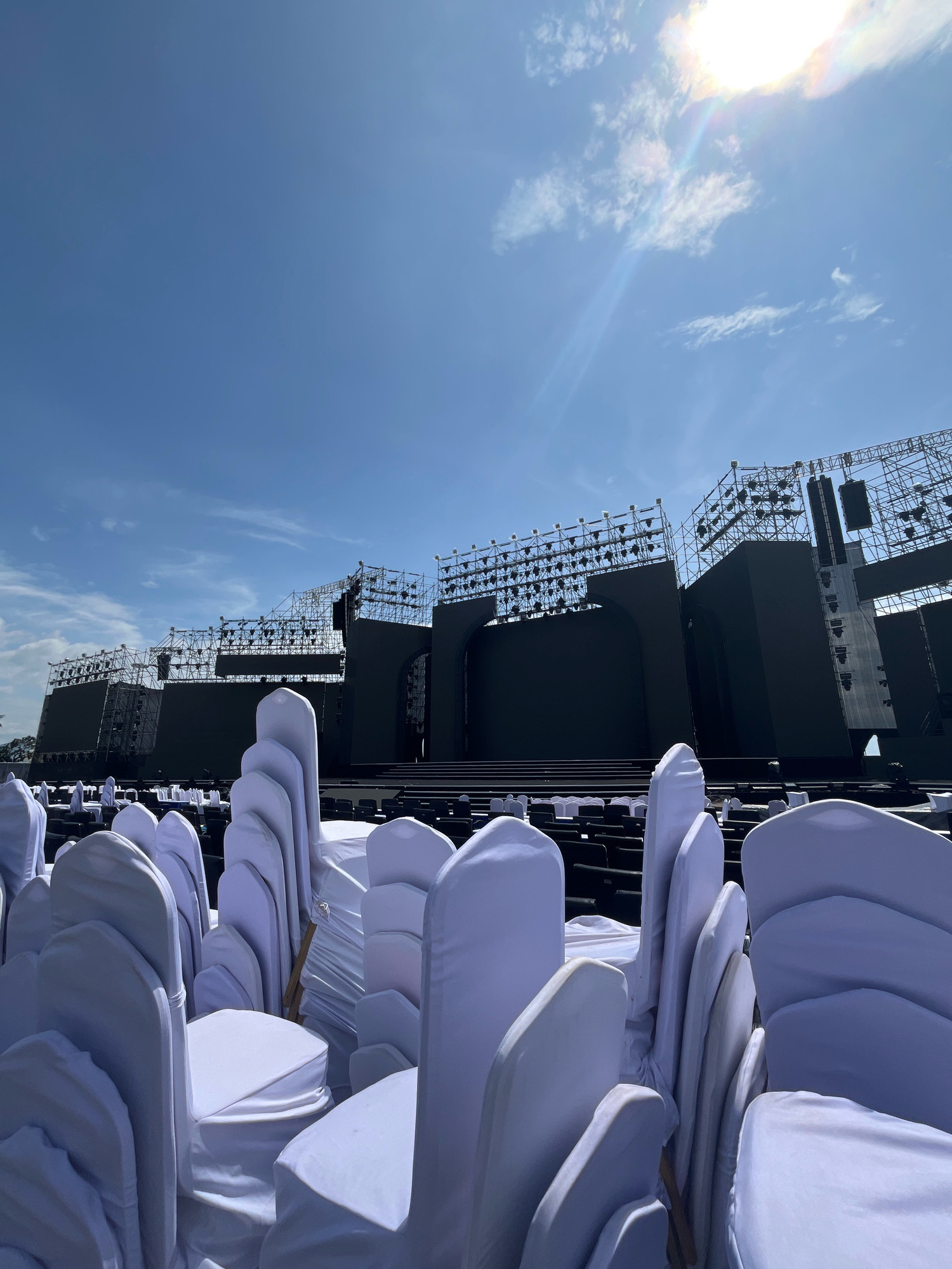 This image captures a wide outdoor stage setup in Nha Trang, Vietnam, under a bright, sunny sky with scattered clouds. In the foreground, rows of chairs are covered in white fabric, creating a neat, almost sculptural appearance. These chairs are arranged in front of the stage, suggesting an event is either about to happen or has recently concluded.
The stage itself is massive, featuring multiple large, dark screens and an intricate scaffolding structure above, laden with numerous lights and speakers. The scale of the stage implies it is designed for a significant performance or event. The background reveals a hazy landscape, possibly indicating proximity to the coast or other urban structures. The overall scene is one of preparedness and anticipation, with the covered chairs hinting at a waiting audience for a spectacle on the grand stage.